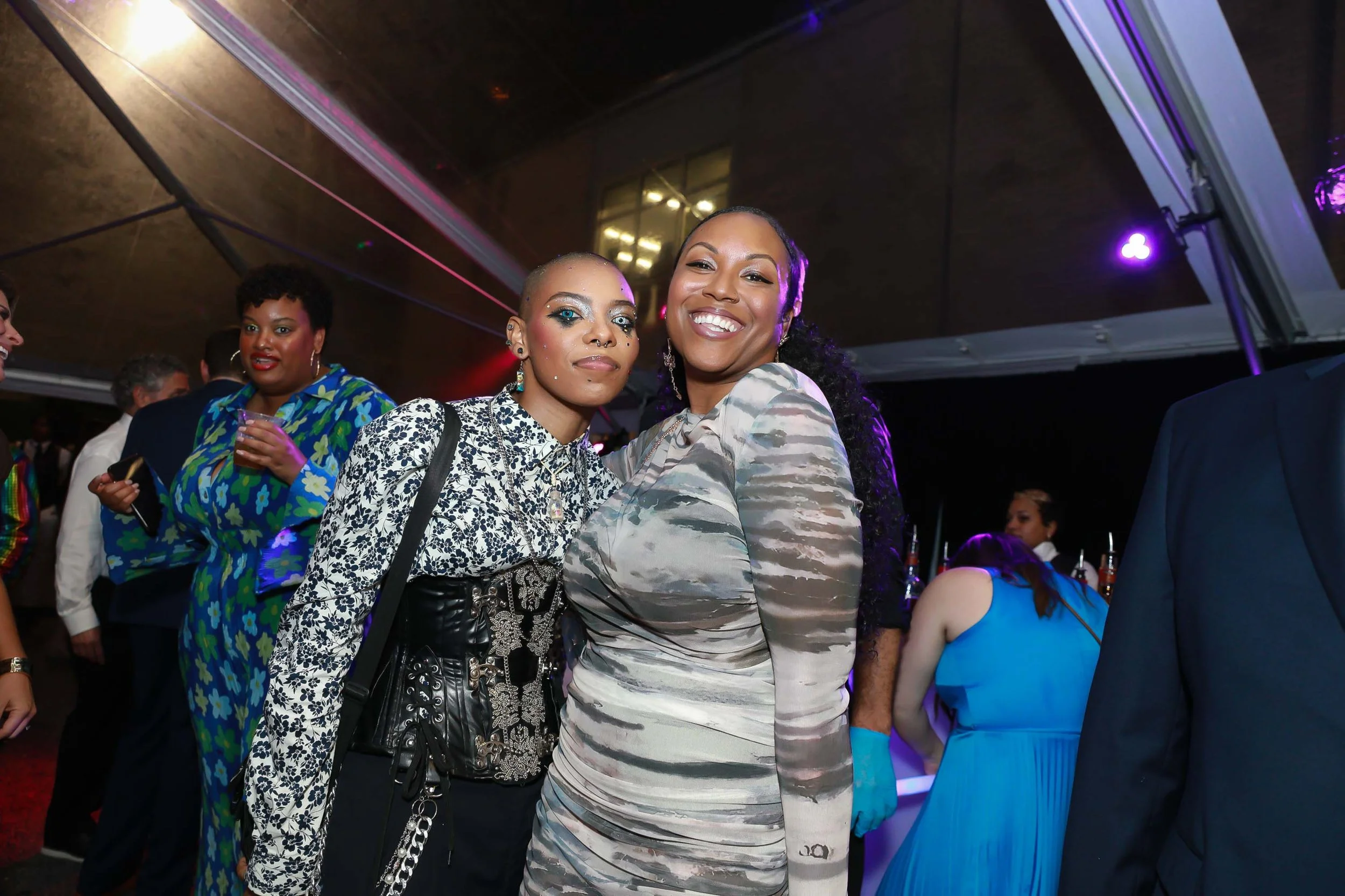 Two women smiling and posing together at a party or event, with other people in the background. One has a short hairstyle with makeup and jewelry, the other has long curly hair and a patterned dress.