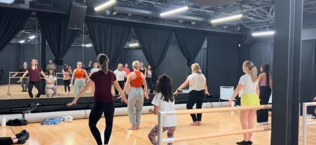Ballet class with students practicing at the barre in front of a mirror with an instructor leading the session.
