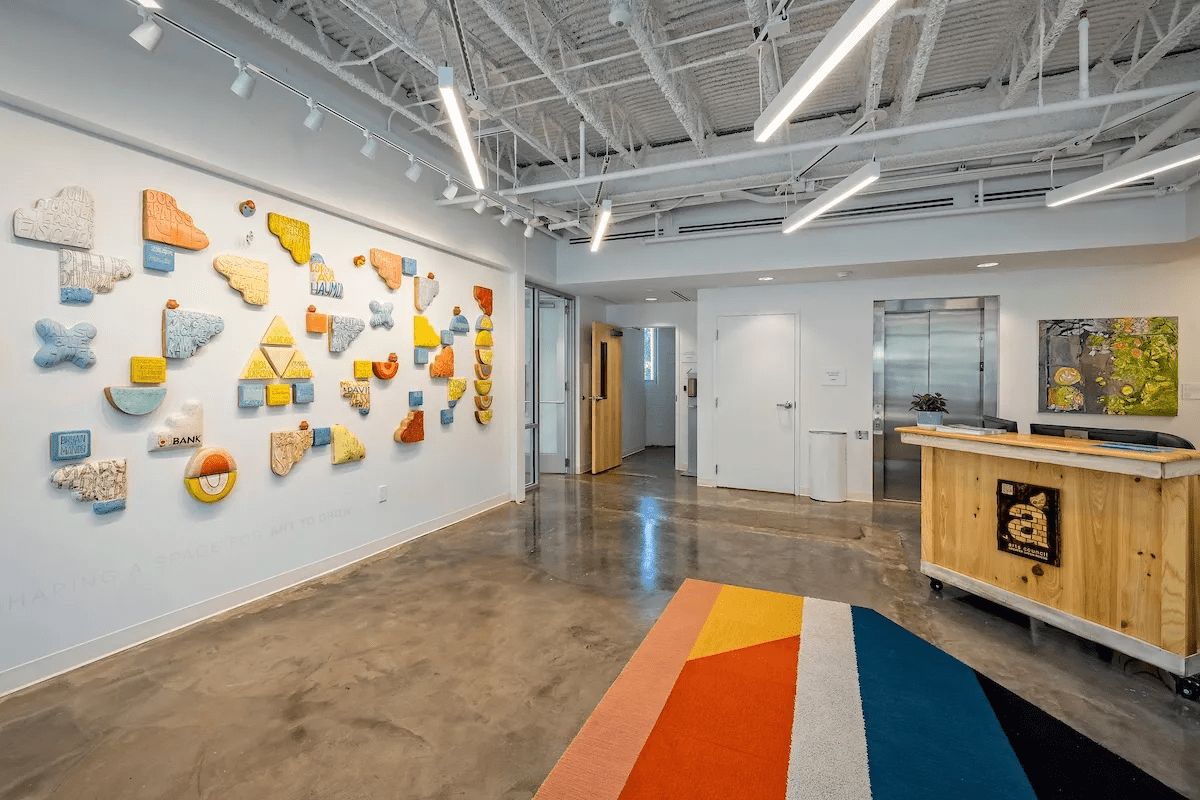 Modern office lobby with abstract colorful wall art, wooden reception desk, elevator, glass door, and a vibrant striped rug on a polished concrete floor.
