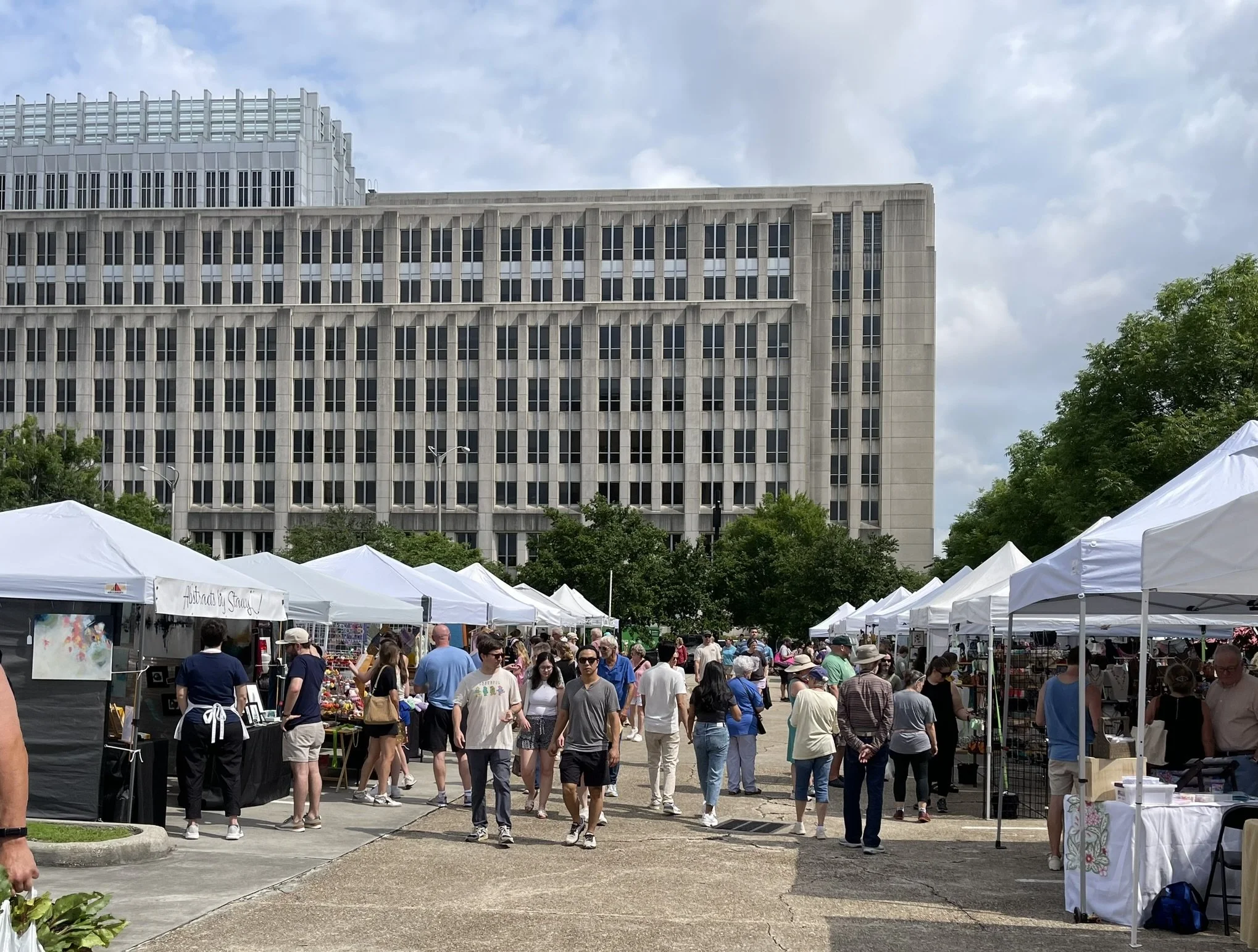 People walking and browsing at an outdoor market with white tents, in front of a tall, modern office building under a partly cloudy sky.