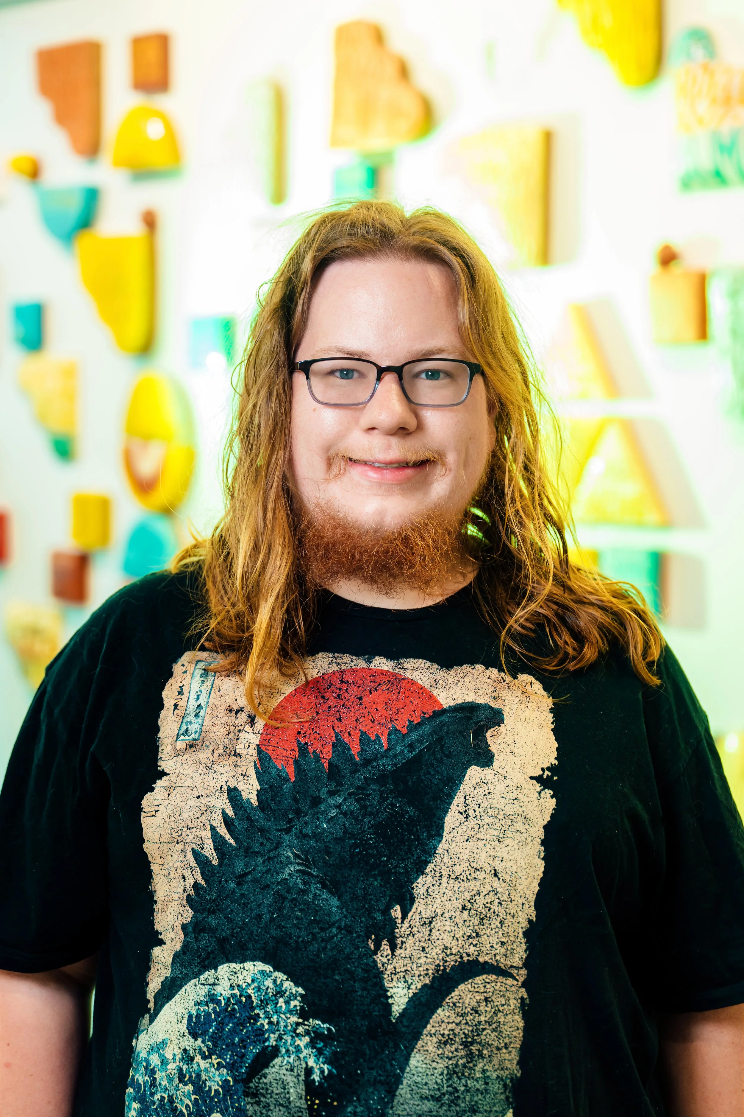 A person with long, wavy red hair wearing black glasses and a black T-shirt with a graphic of a skull and a red, white, and blue design.