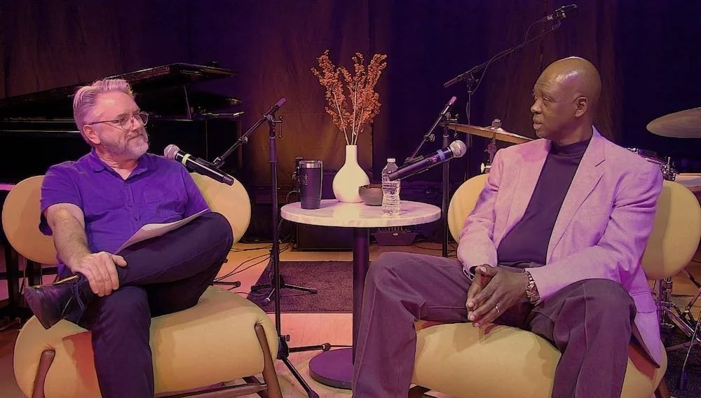 Two men having a conversation during a podcast or interview in a music studio with microphones, a piano, and musical instruments in the background.