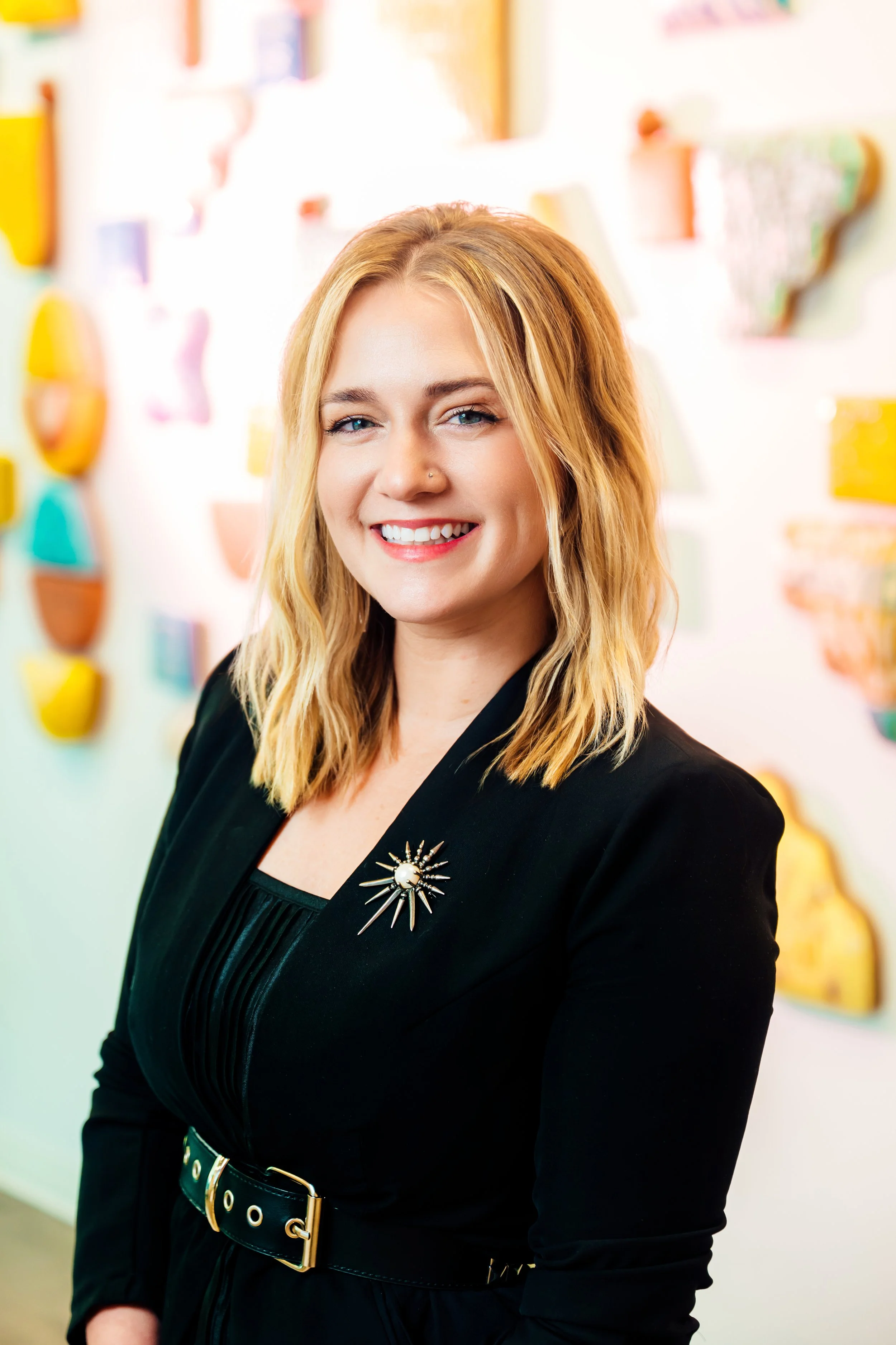 A smiling woman with blonde hair and blue eyes, wearing a black blazer with a brooch and standing in front of a colorful, blurred background.