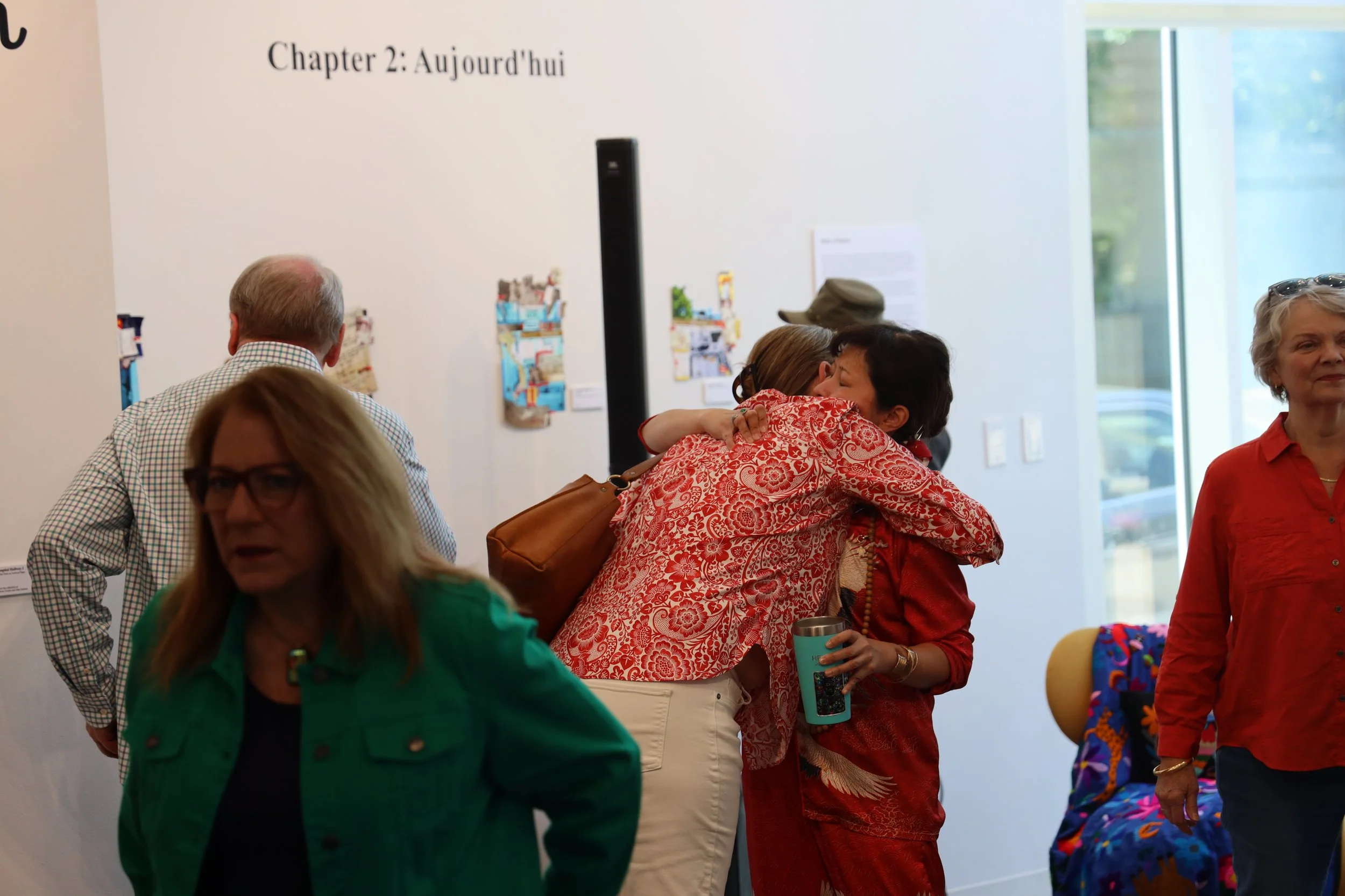 People gather at an indoor art exhibit, with colorful artwork displayed on white walls, and sunlight streaming through large windows.