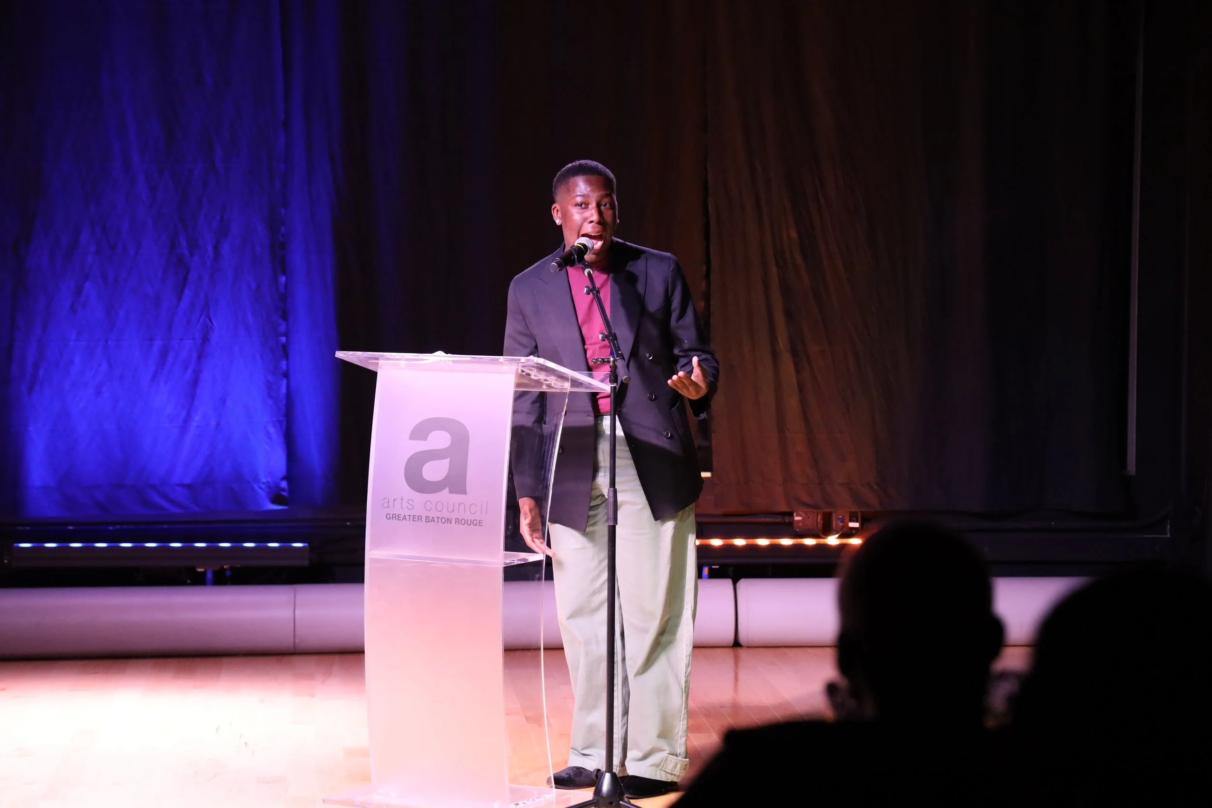 A person speaking at a podium during an event with a sign reading "arts council GREATER BATON ROUGE". The person is wearing a dark blazer, burgundy shirt, and light-colored pants, and they are standing on a stage with blue and brown curtains.