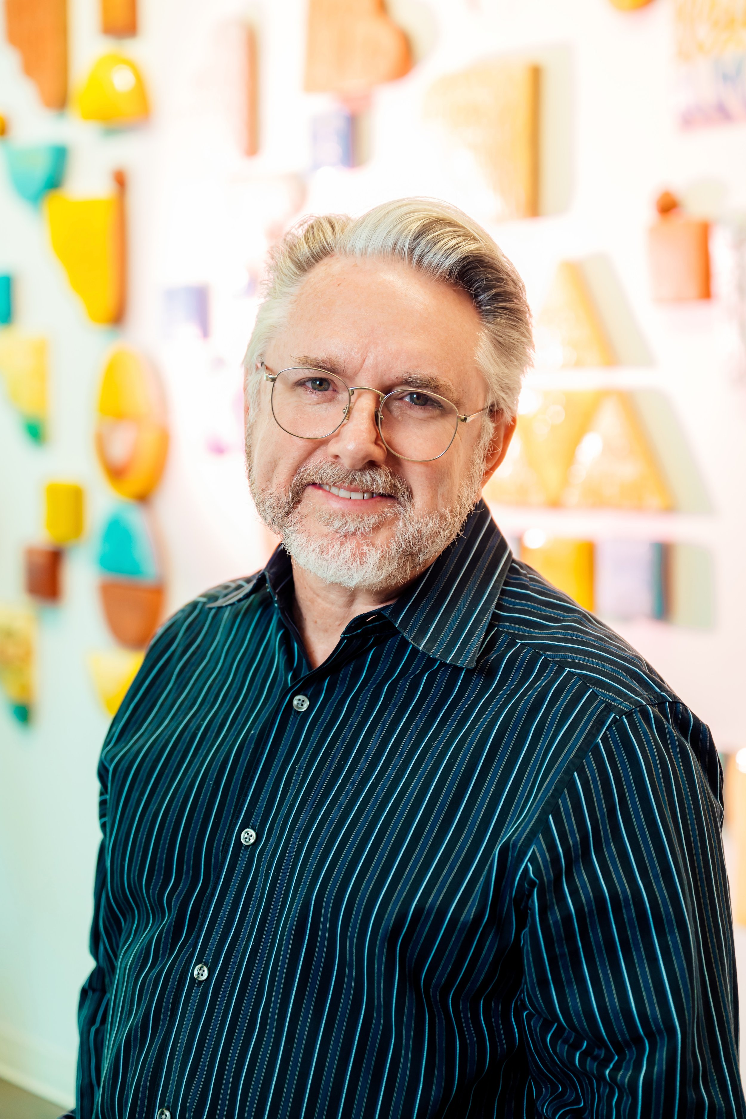 A smiling older man with gray hair, wearing glasses and a dark blue striped shirt, standing in front of a colorful wall with various geometric shapes and art pieces.