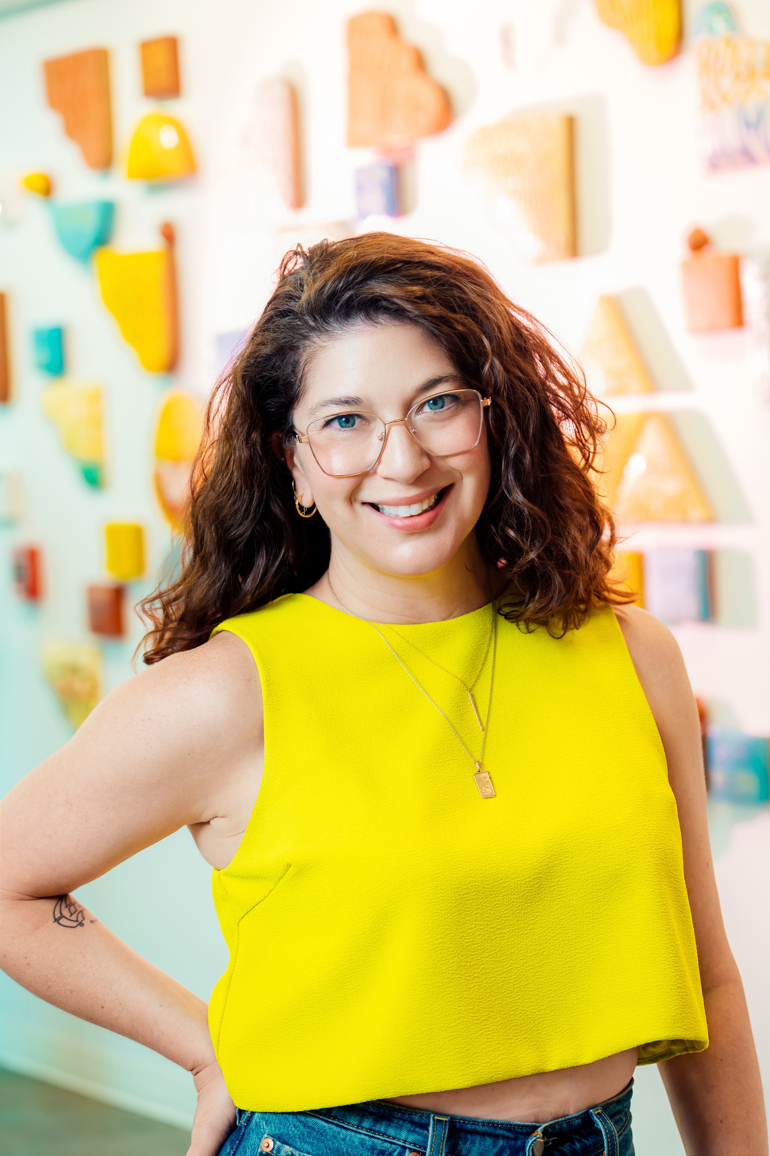 A woman with curly brown hair, wearing glasses and gold hoop earrings, is smiling and posing with one hand on her hip. She is wearing a bright yellow sleeveless top and blue jeans. The background features colorful abstract wall decorations.