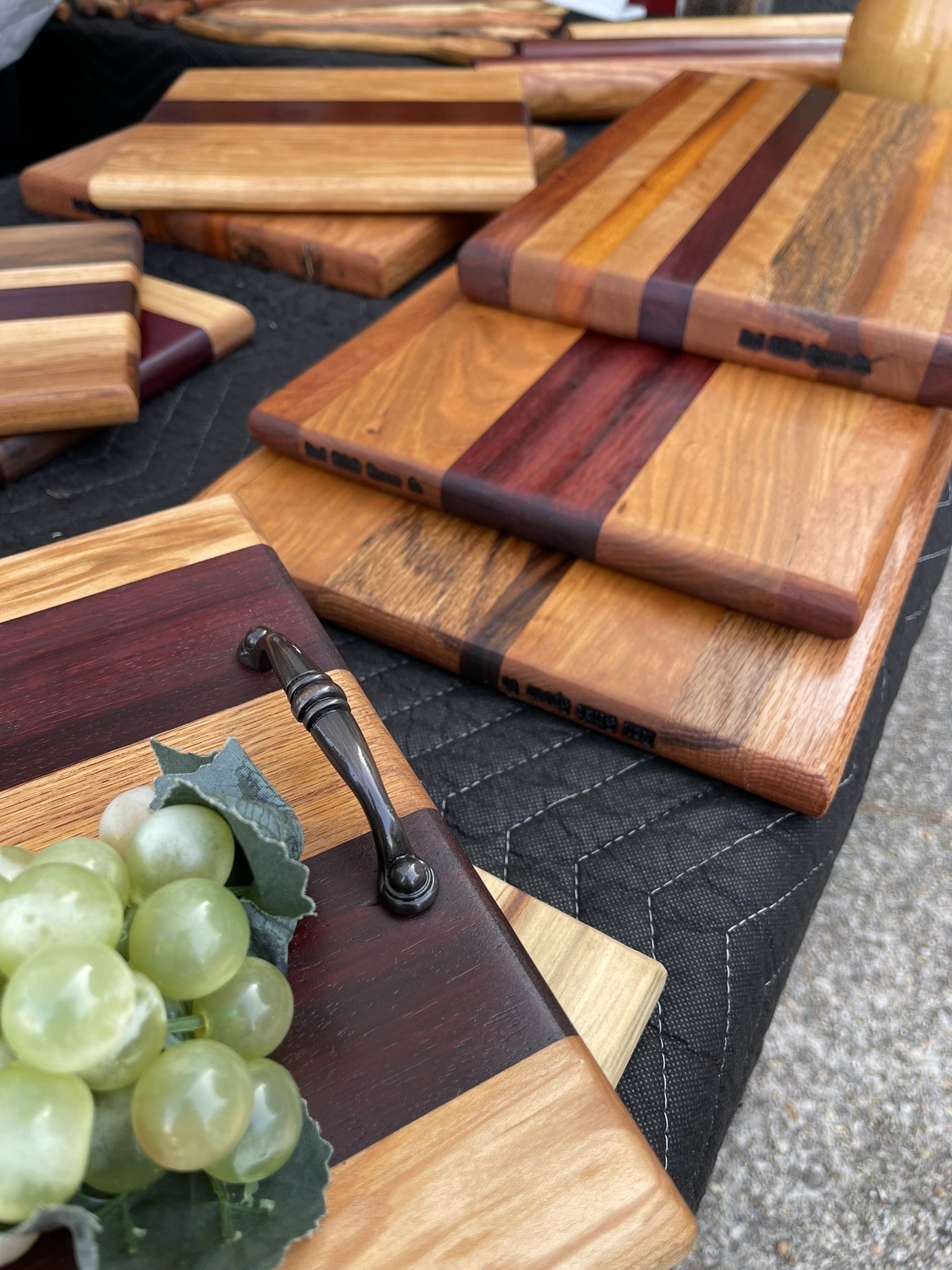 Multiple wooden cutting boards with various wood grains and colors arranged on a black fabric surface, with a bunch of green grapes in the foreground.