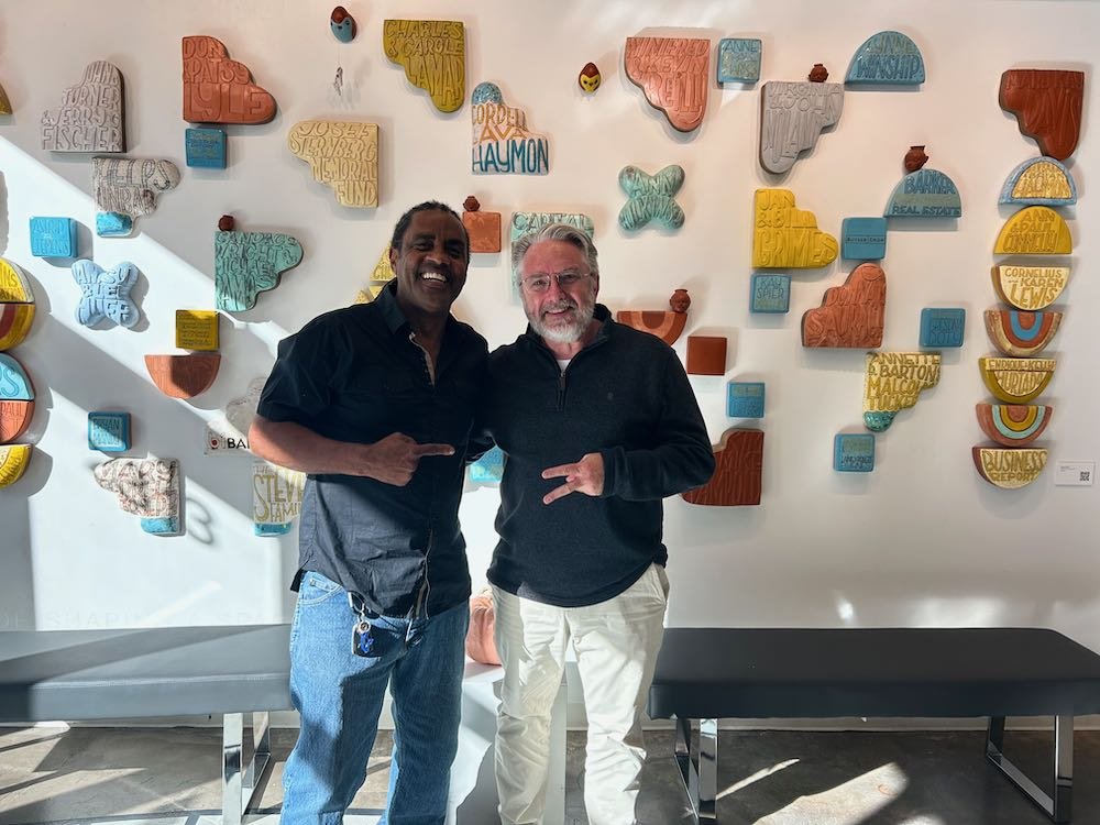 Two men smiling and posing in front of a wall with colorful, three-dimensional map-like wall art.