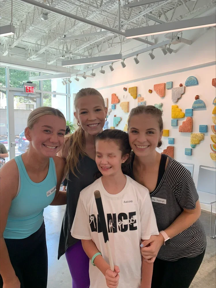 Four smiling young women and a girl with a bandage on her face inside an art gallery or studio, with colorful textured wall art in the background.