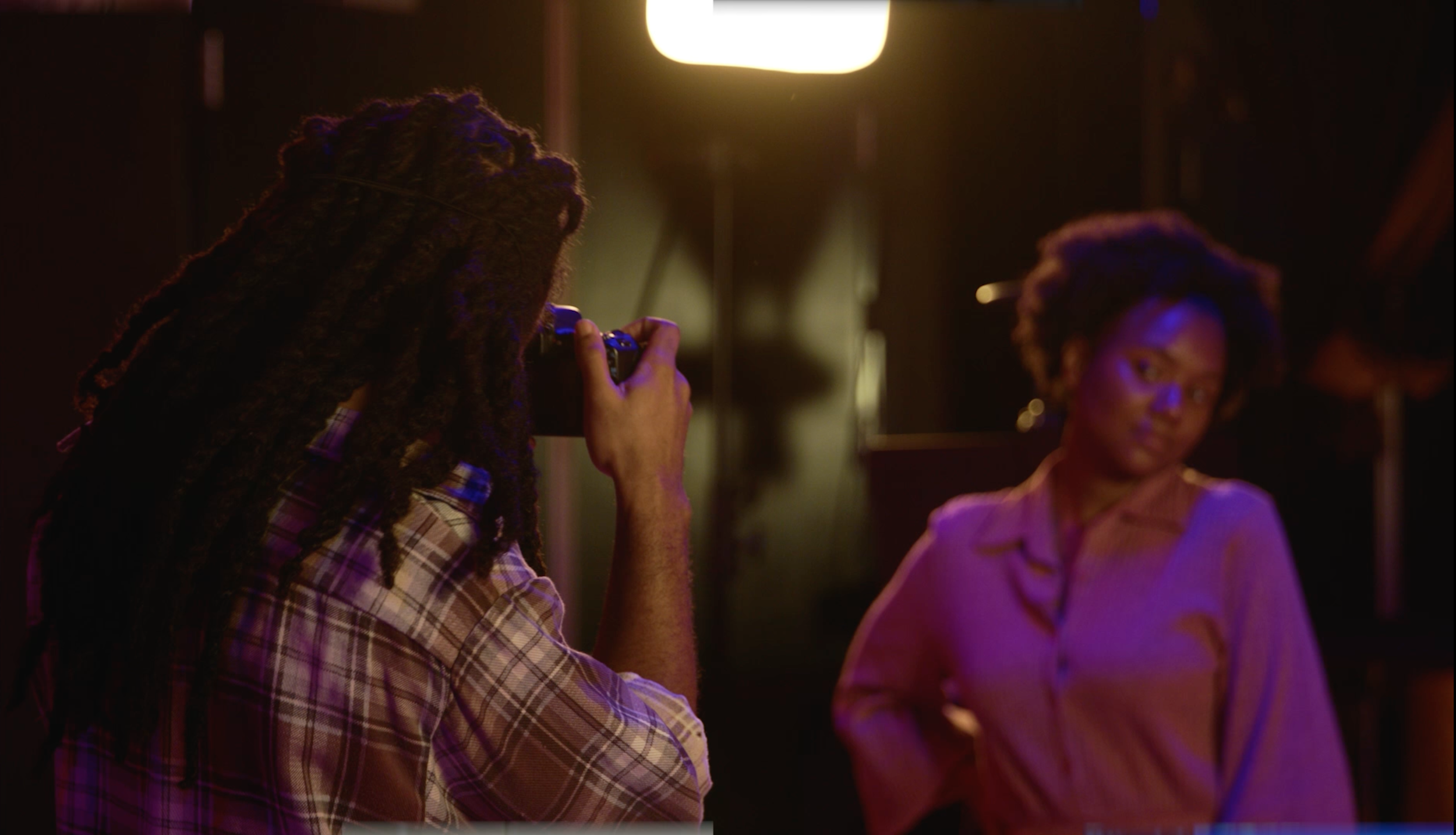 A person with long dreadlocks in plaid shirt taking a photo of a woman in a pink top in a dimly lit room.