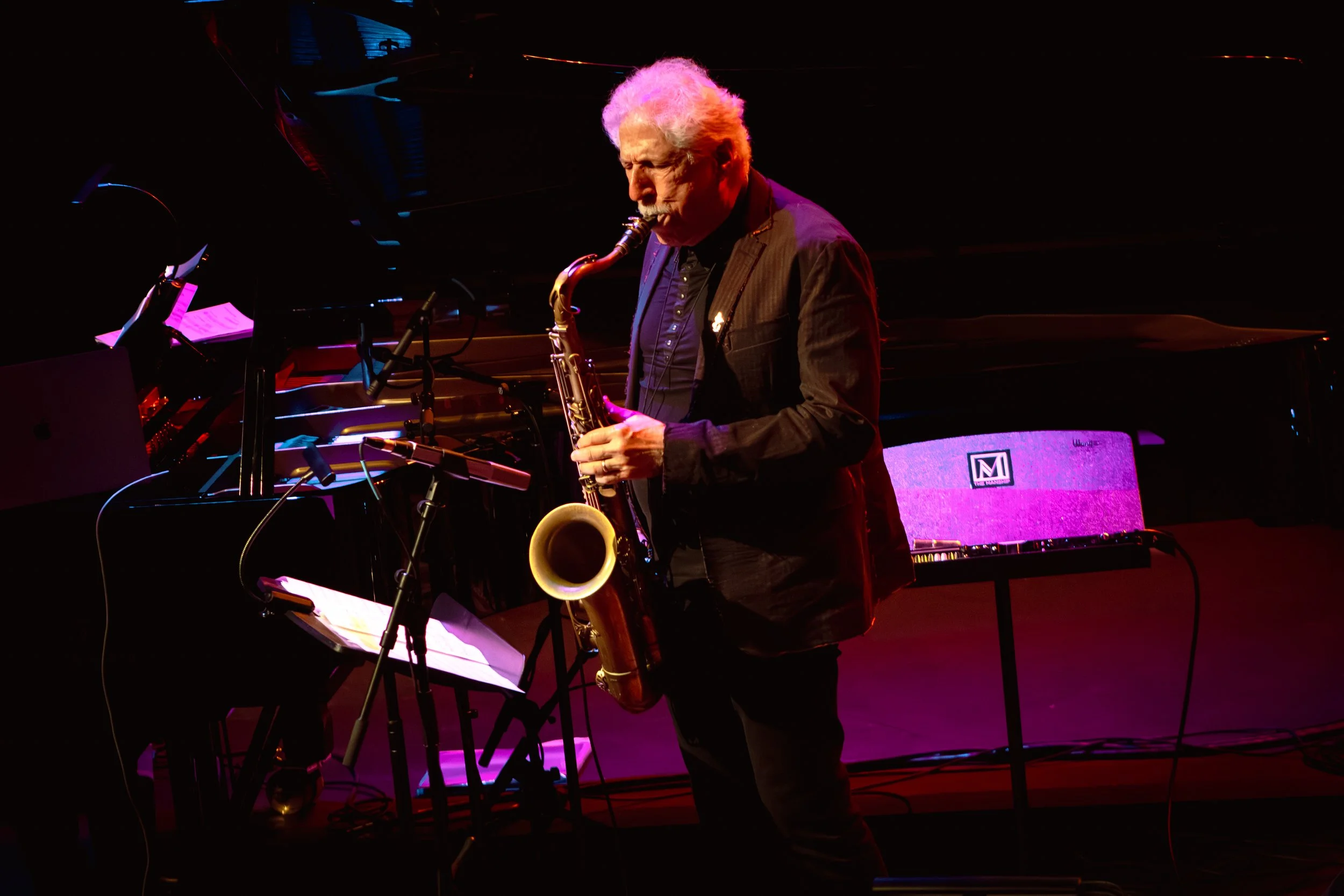 An elderly man with gray hair and a gray beard playing a saxophone on stage. He is wearing a dark suit and is standing next to a black grand piano with sheet music and various microphones nearby. The stage is lit with pink and purple lighting, creating a vibrant atmosphere.