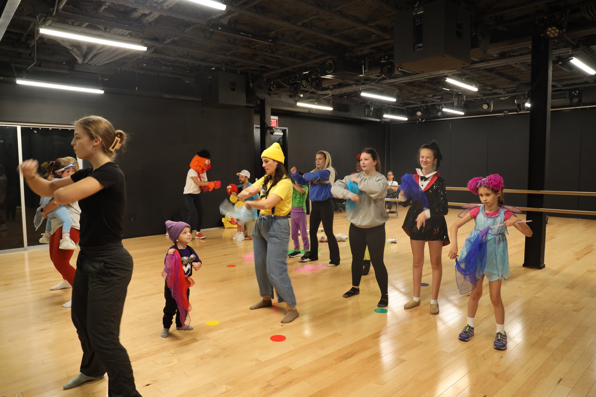Children and adults in a dance studio dressed in colorful costumes, some wearing hats or accessories, participating in a dance or theatre activity.