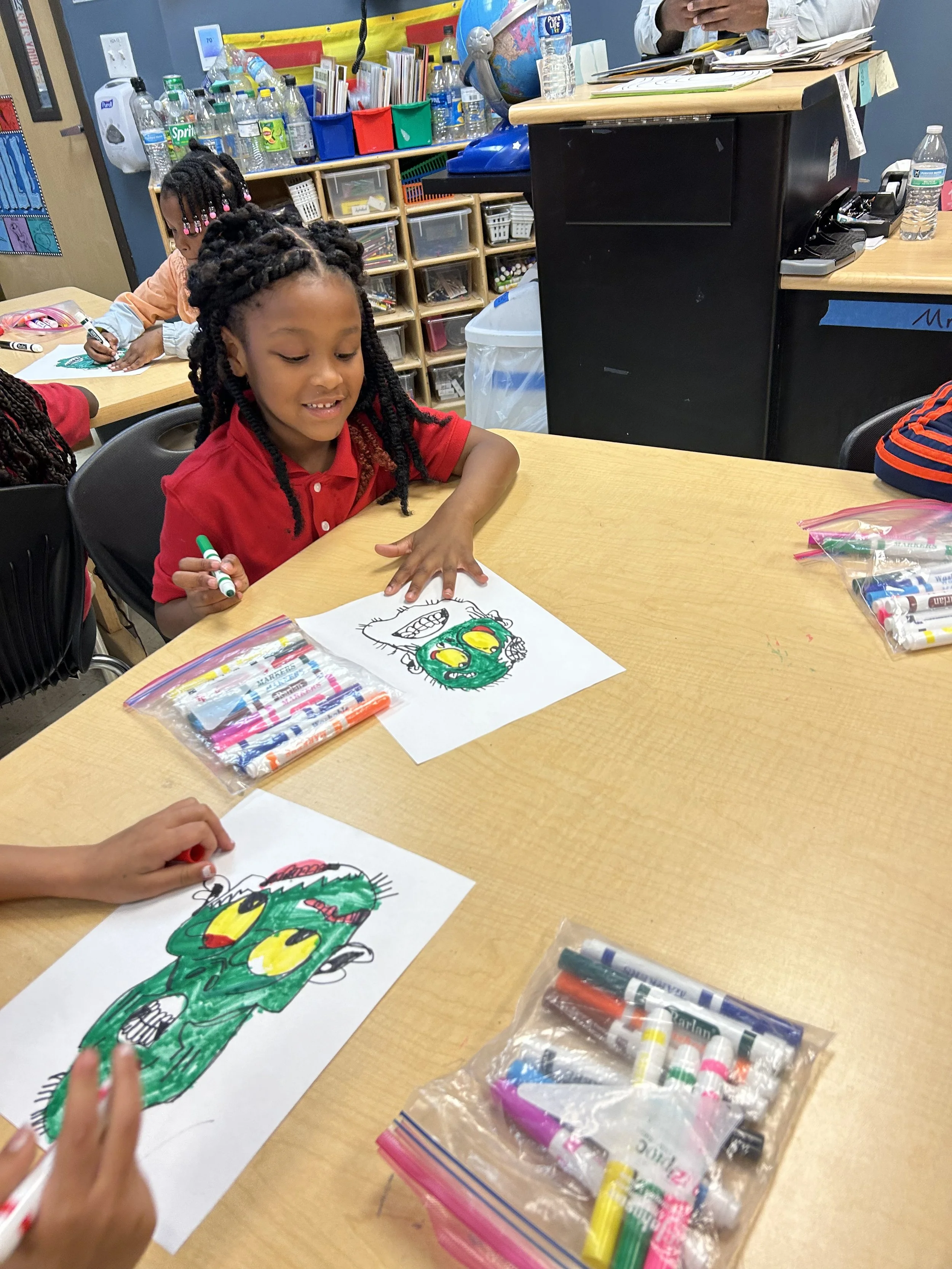Children sitting at a table, coloring pictures of cartoonish green monsters with yellow eyes and red mouths using markers. The table holds bags of markers and coloring supplies, with a classroom bookshelf and supplies visible in the background.