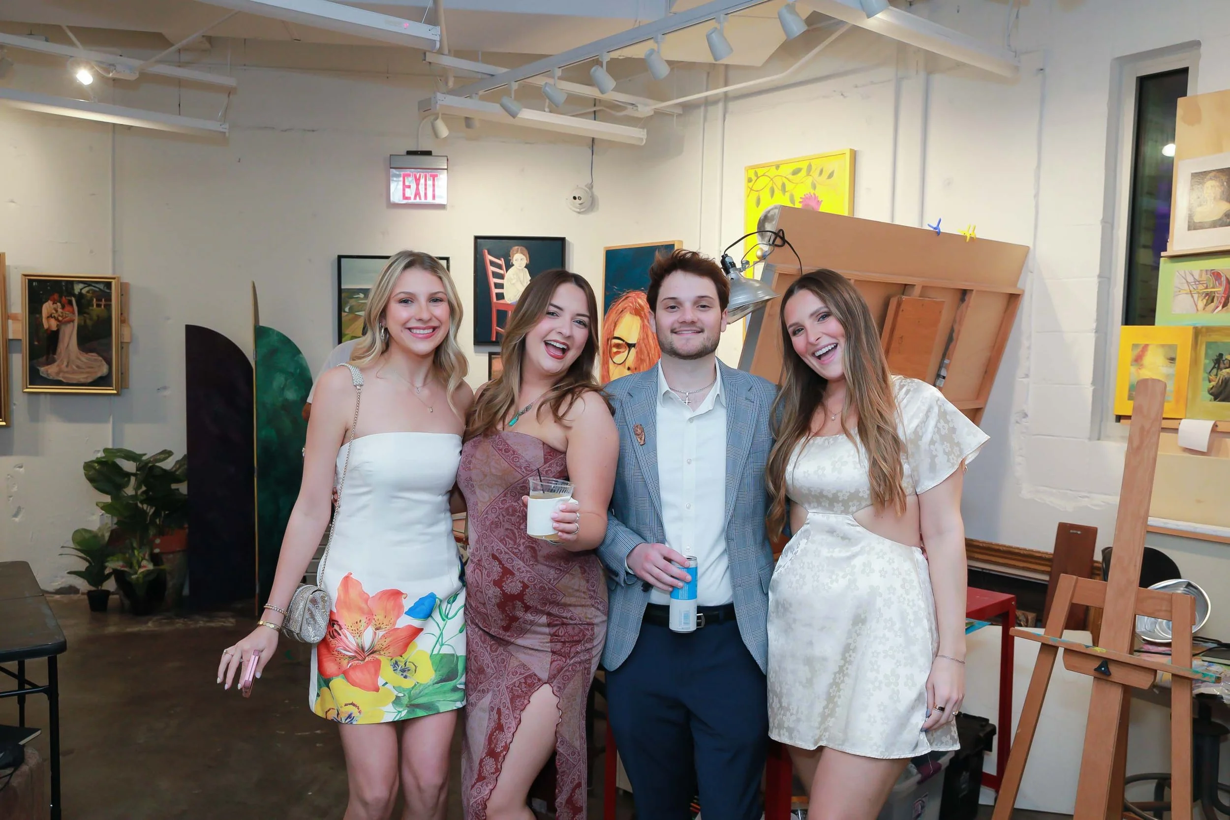 Four young adults, three women and one man, are smiling and posing together in an art gallery or studio, surrounded by various paintings and artwork on the walls.
