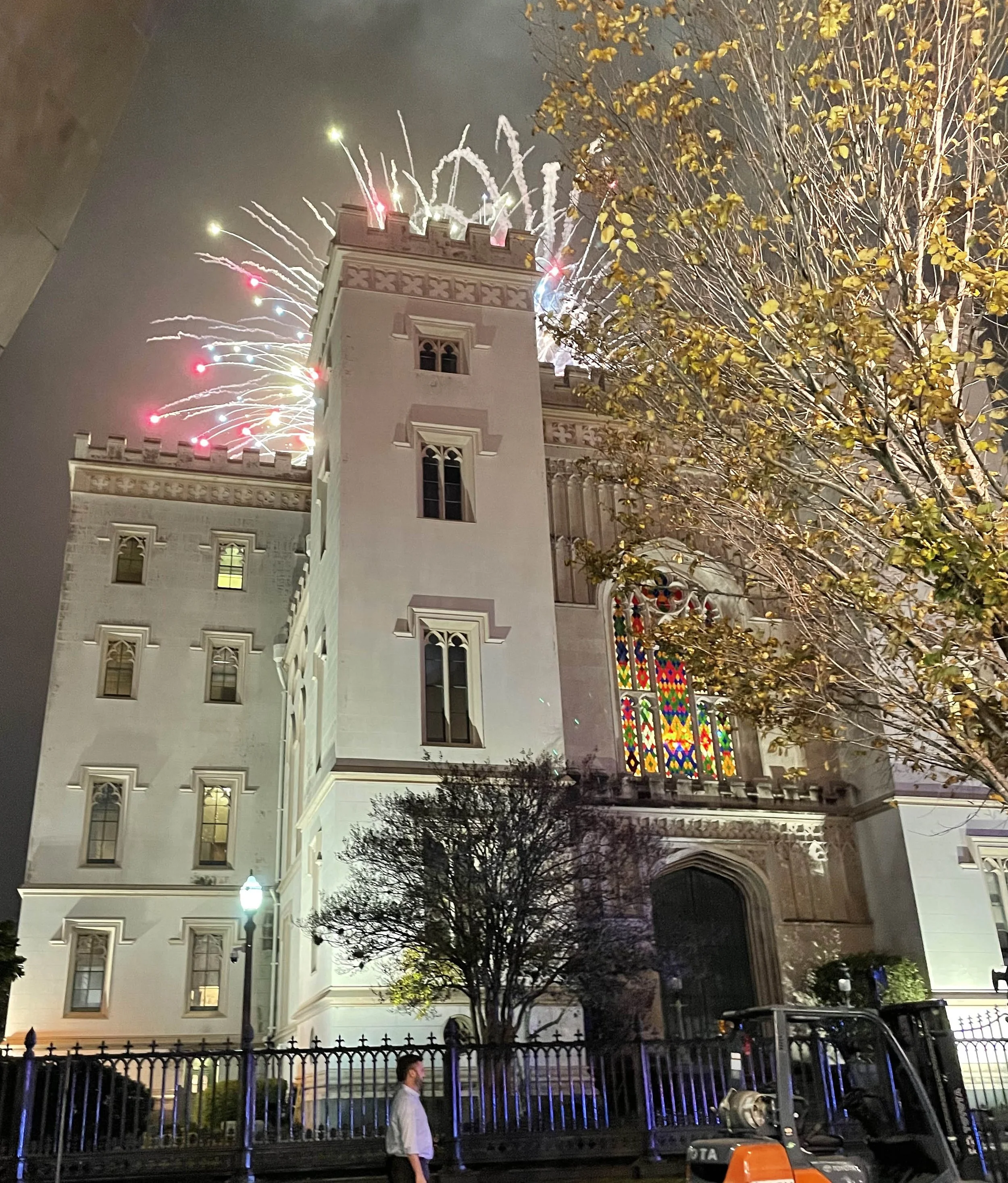 Night view of a white castle-like building with arched windows, fireworks lighting up the sky behind it, and red and yellow leaves on a tree on the right side of the image. There are streetlights and a person walking near a fence in the foreground.