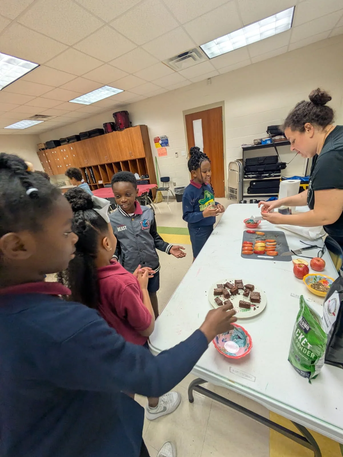 A group of children gather around a table in a room, watching a woman prepare snacks with apples and brownies, in an educational or community setting.