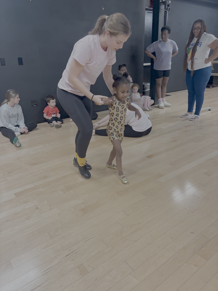 A young woman with brown hair in a ponytail, wearing a white T-shirt and black pants, is helping a young girl in a leopard print dress practice dance steps in a studio with a wooden floor. Several other children and adults are watching, some sitting on the floor, in front of a dark wall.