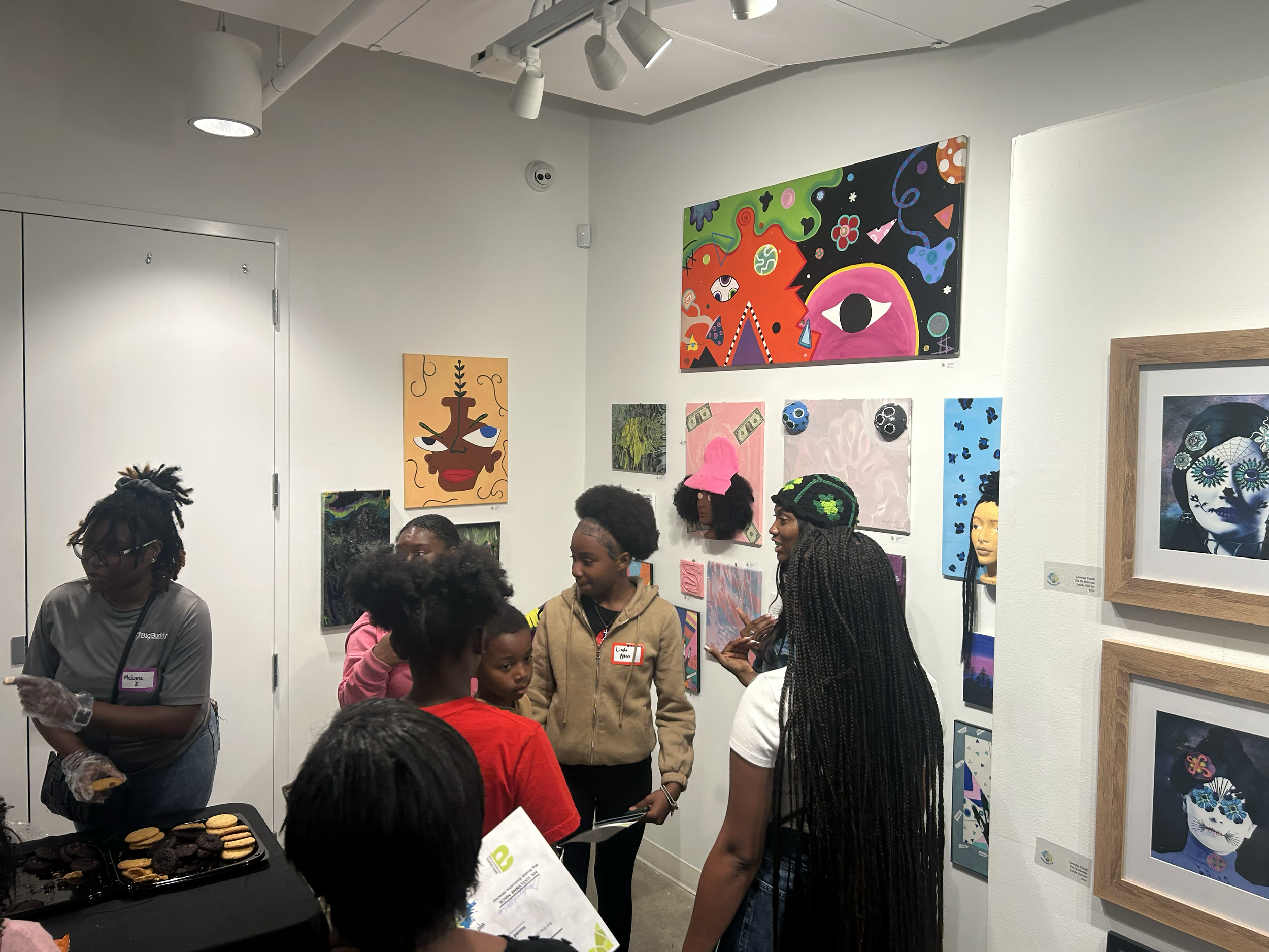 People gathered in an art gallery, viewing various colorful artworks on the walls, with a tray of assorted cookies on a table in the foreground.