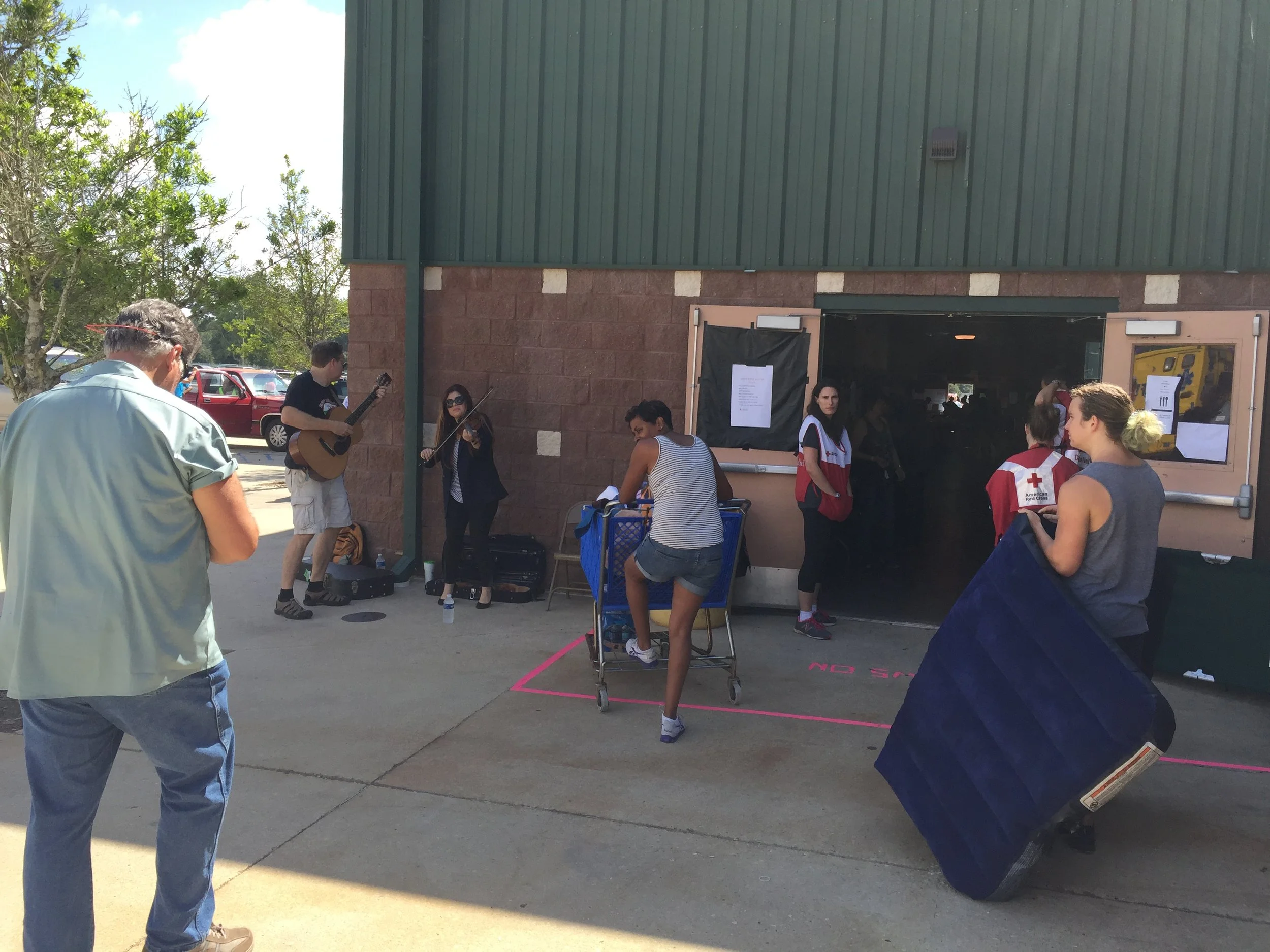 People gathered outside a building, with some playing musical instruments and others waiting in line or talking, during daytime with clear skies.