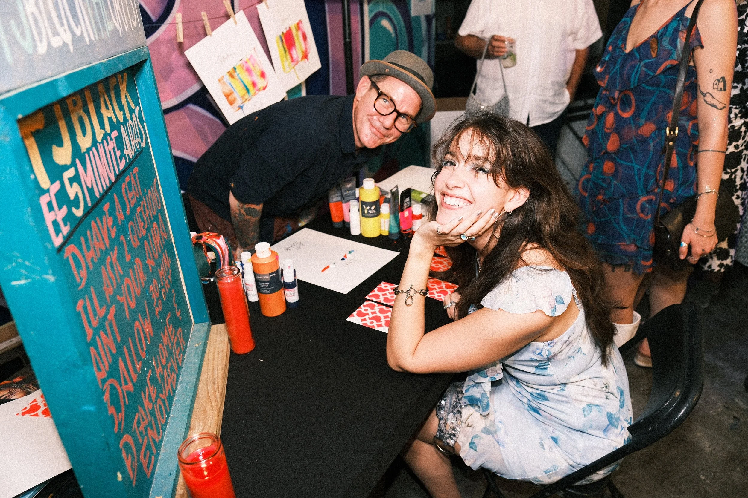A woman with curly brown hair and a light blue dress is sitting at a table, smiling, with her hand resting on her chin. A man with glasses, a hat, and tattoos on his arms is leaning over the table, smiling at the camera. The table has art supplies including paint bottles, and there is colorful artwork on the wall behind them, along with a sign for a festival or event.