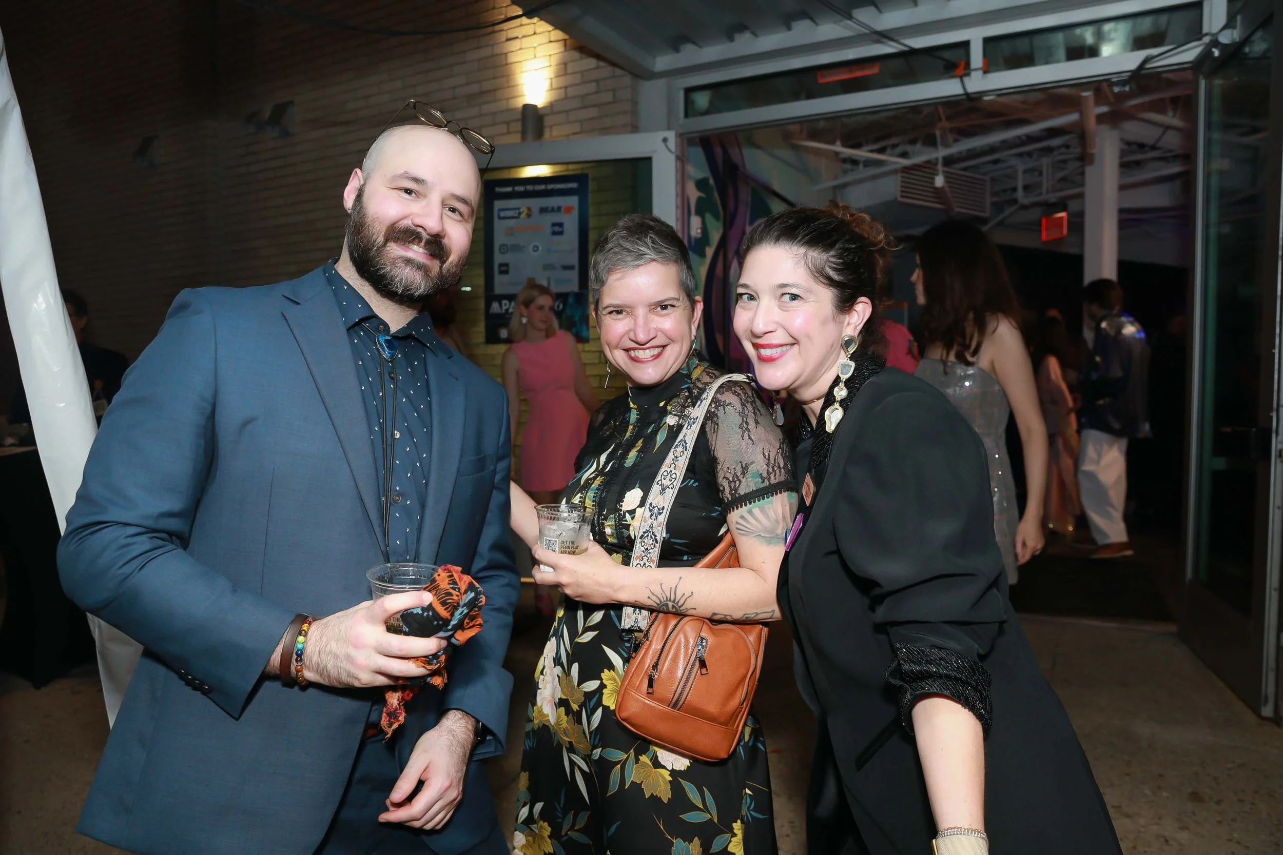 Three people smiling at an indoor event, two women and one man, dressed in stylish clothing, holding drinks, with a background of other attendees and colorful wall art.
