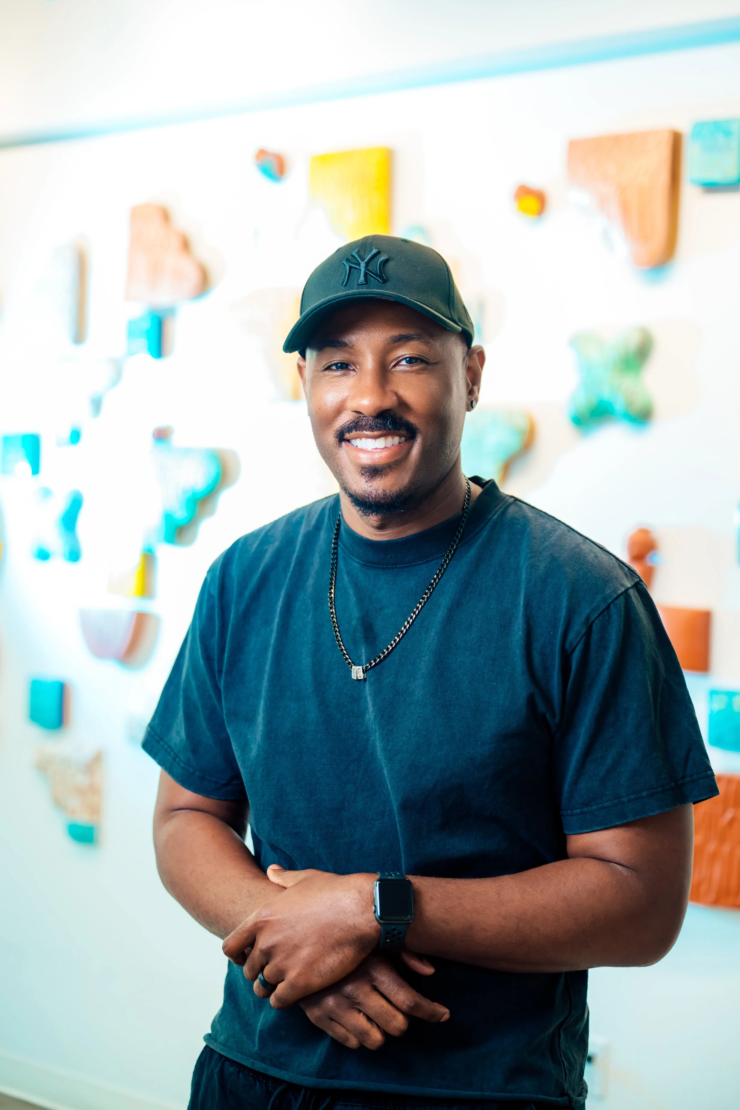 A smiling man wearing a black T-shirt, a black cap with a New York Yankees logo, a wristwatch, and a necklace, standing in front of a colorful abstract wall art.
