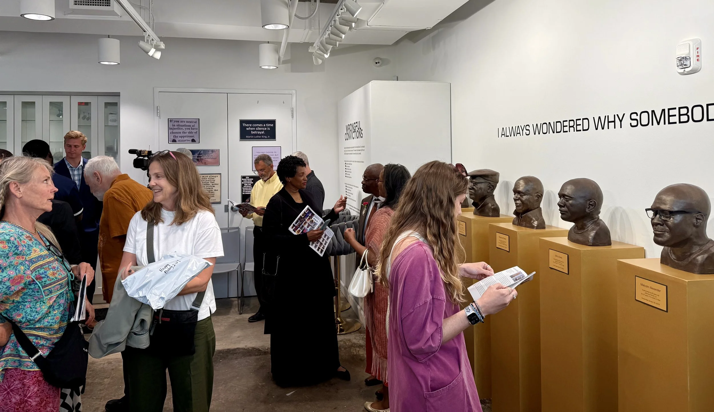A group of people inside a museum or gallery, viewing bust sculptures of various historical figures placed on yellow display stands. Some people are reading pamphlets, and informational posters are visible on the wall.