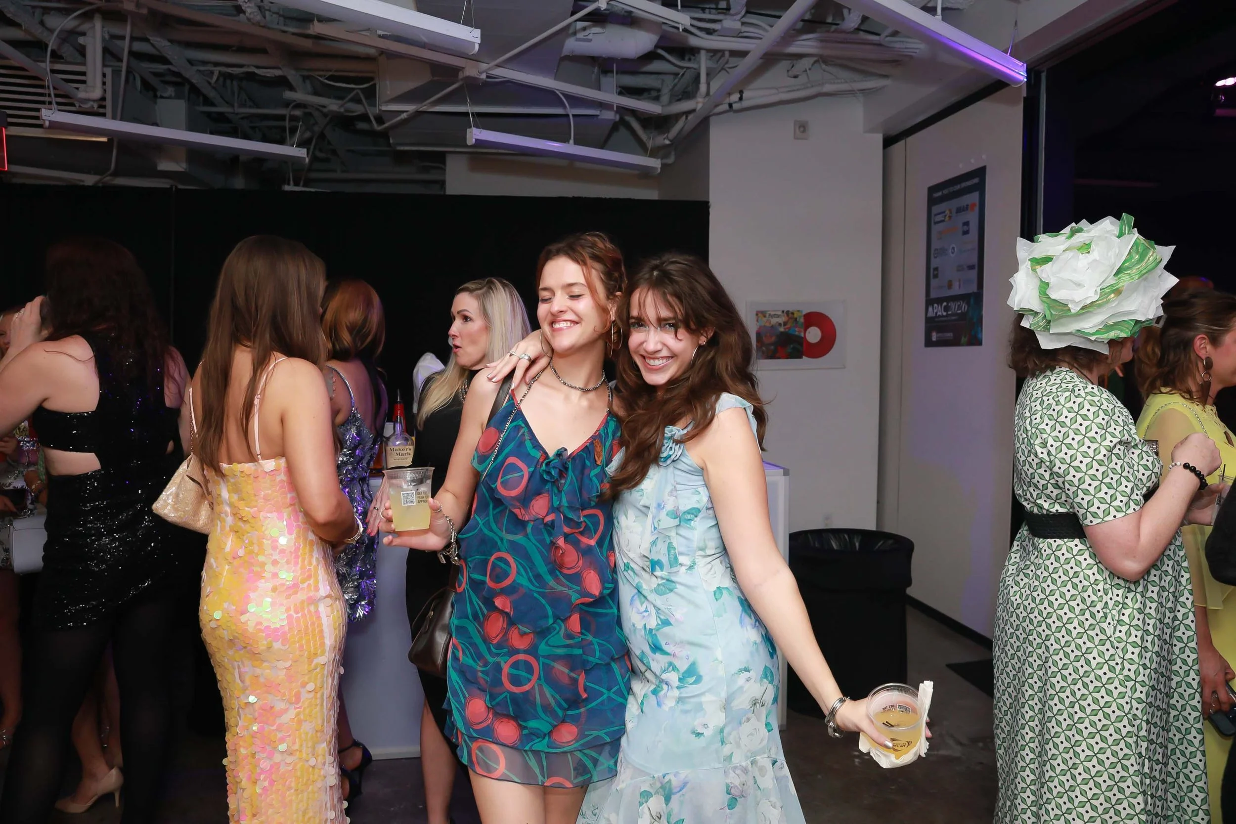 Joyful women at a party, two smiling women with drinks in hand in the foreground, surrounded by others socializing in a festive setting with colorful clothing and a vibrant atmosphere.