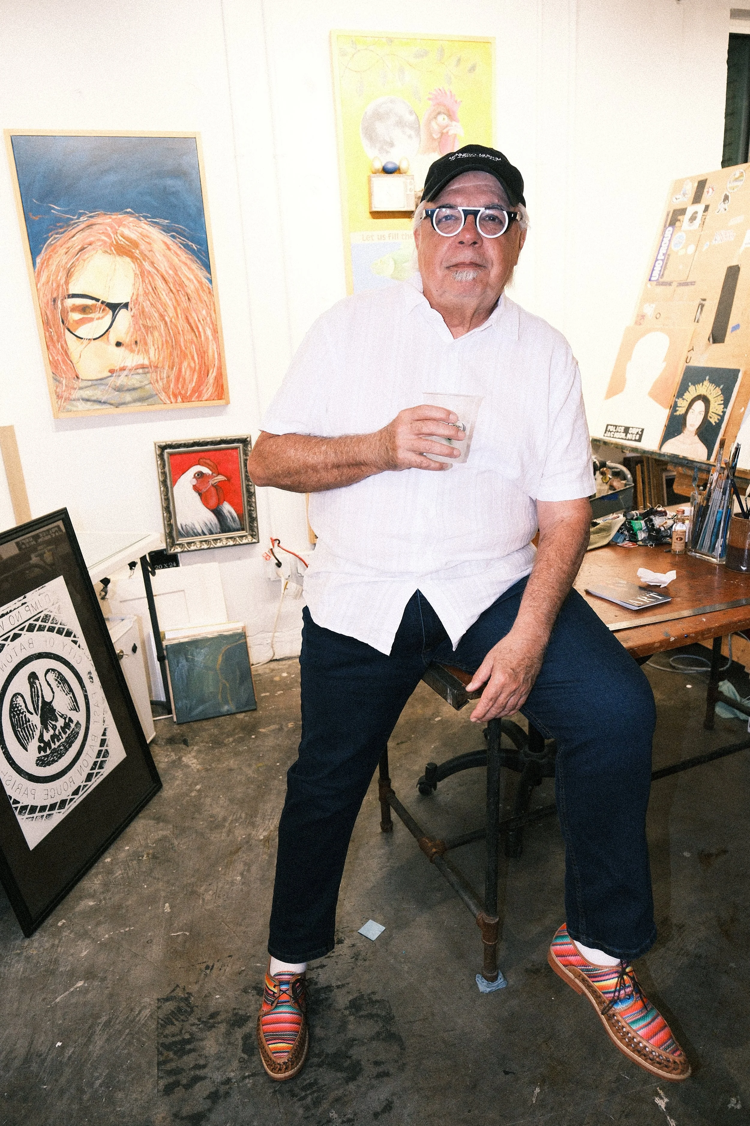 An older man with glasses, a black cap, and a white shirt sitting on a stool in an art studio. The studio walls display paintings of a woman with orange hair, a chicken, and other artwork. The man is holding a drink and wearing colorful striped shoes.