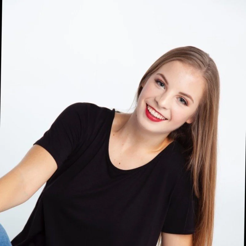 Smiling young woman with long blonde hair wearing a black t-shirt against a plain white background.