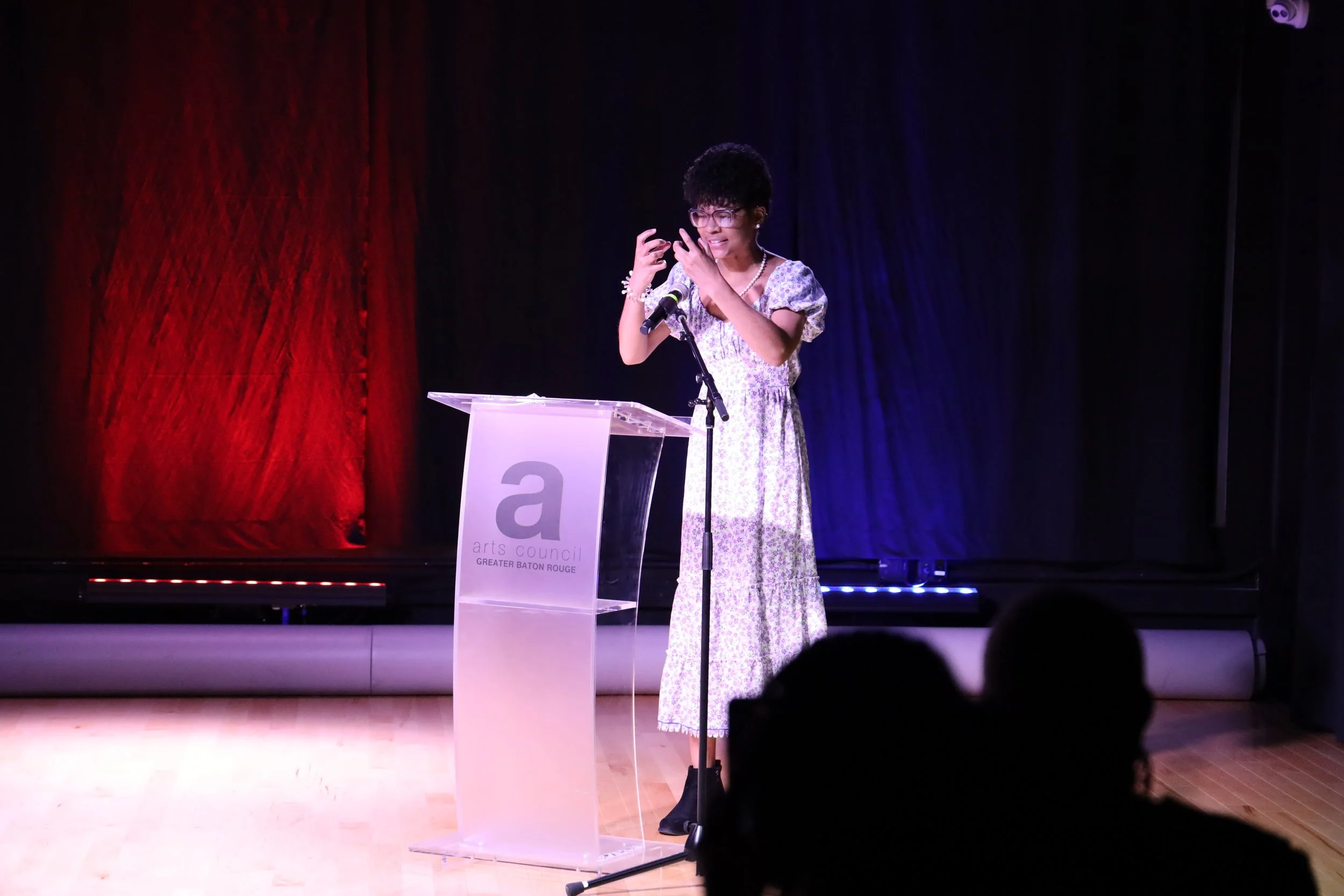 A woman with short curly hair and glasses, wearing a floral dress, stands at a clear podium with a microphone, speaking or presenting on stage with a red and black curtain background.