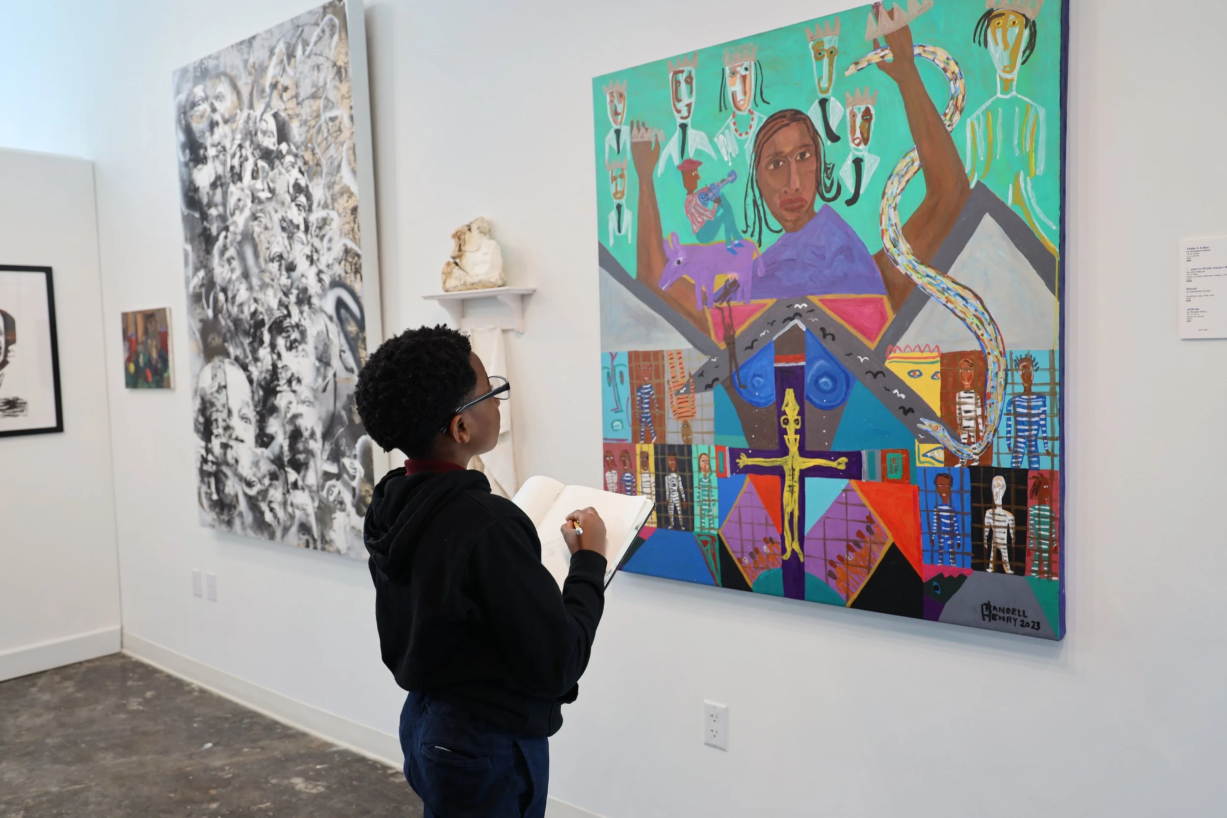 A person with curly hair and glasses standing in front of a colorful abstract painting in an art gallery.