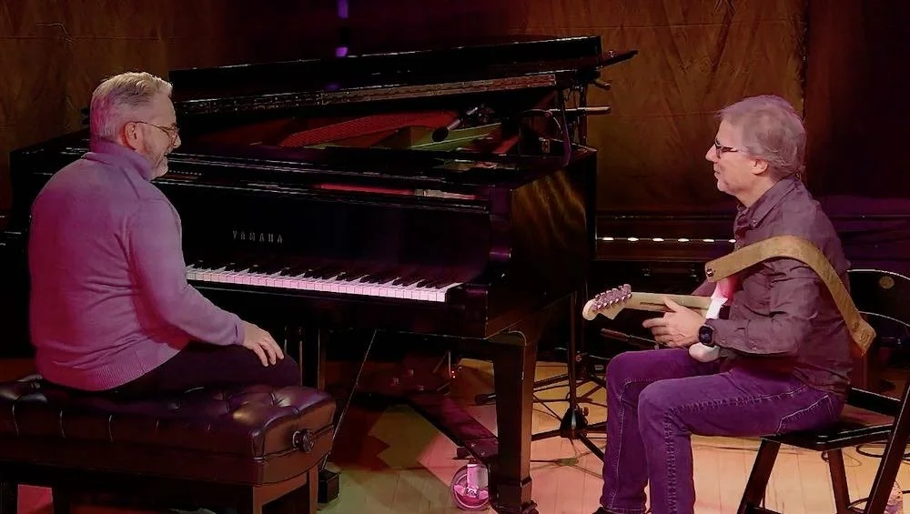 Two men playing musical instruments: one is seated at a Yamaha grand piano and the other is sitting with an electric guitar. They are in a music rehearsal or performance setting.