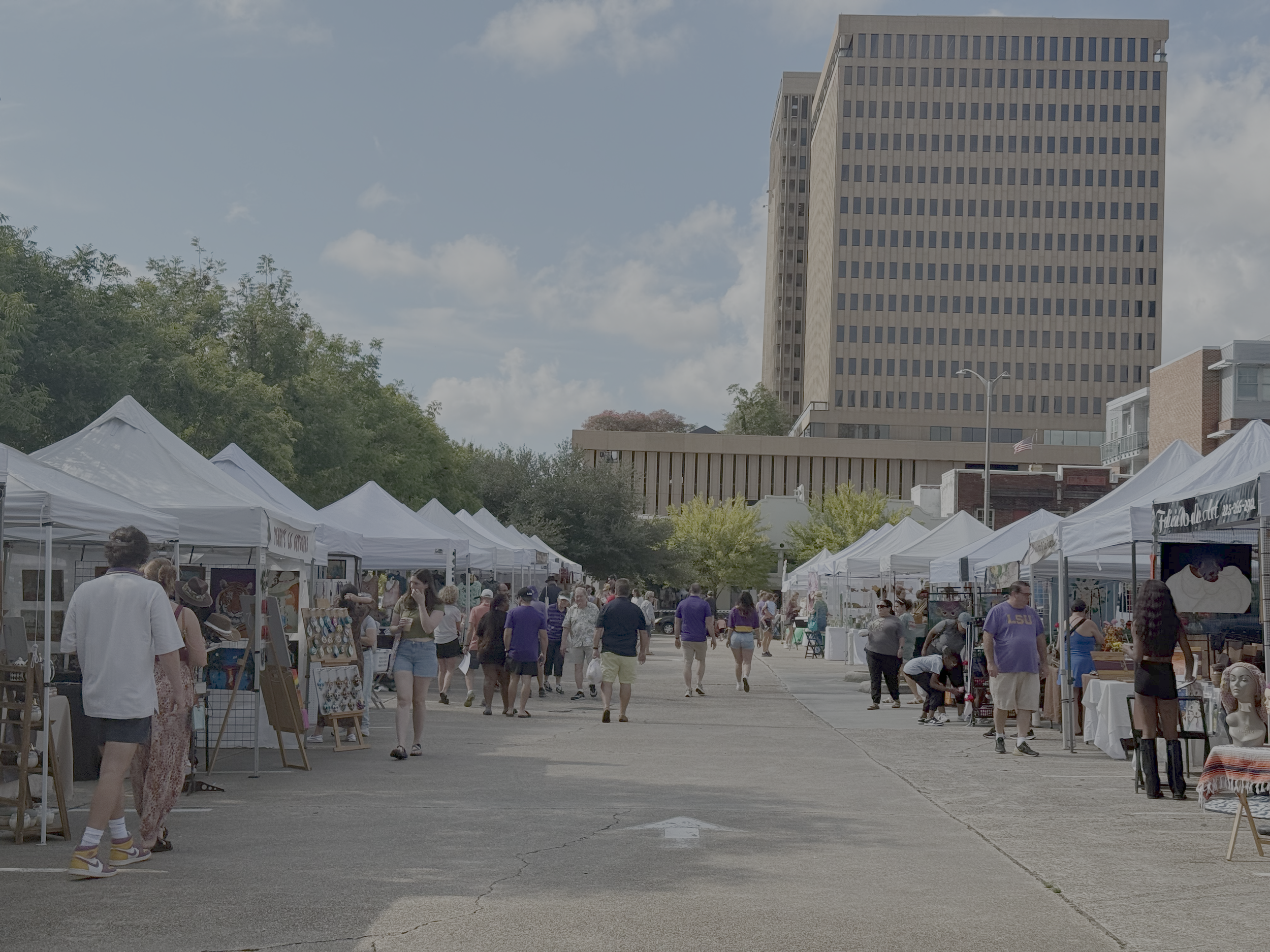 Outdoor market with white tents and people browsing, large office building in the background, partly cloudy sky.