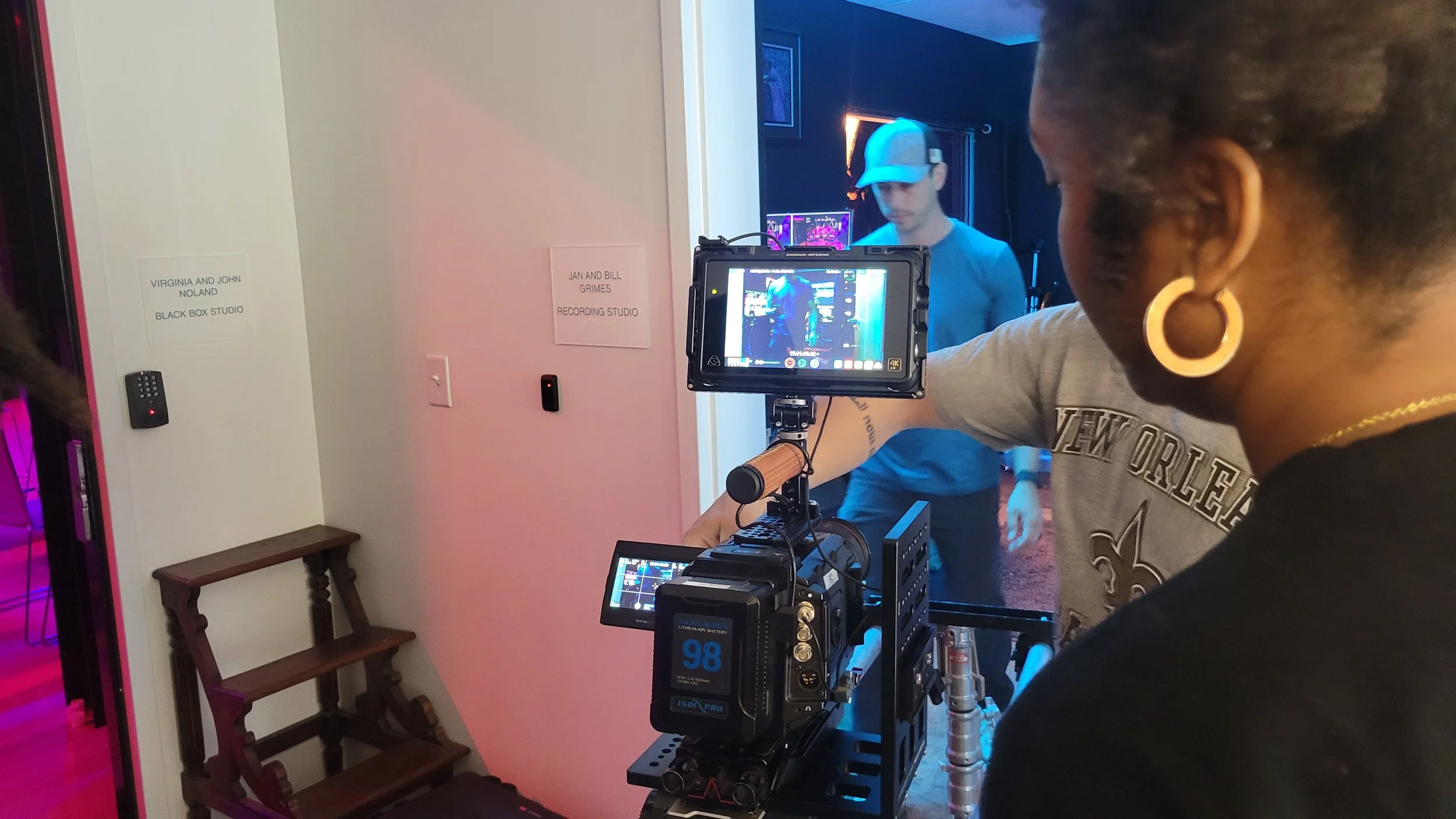 Scene from a film or video shoot showing a woman with dark hair in hoop earrings operating a camera, recording a young man in a blue shirt and cap in a studio room with signs indicating 'Virginia and John Noland, Black Box Studio' and 'Jan and Bill Grimes, Recording Studio' on the wall.