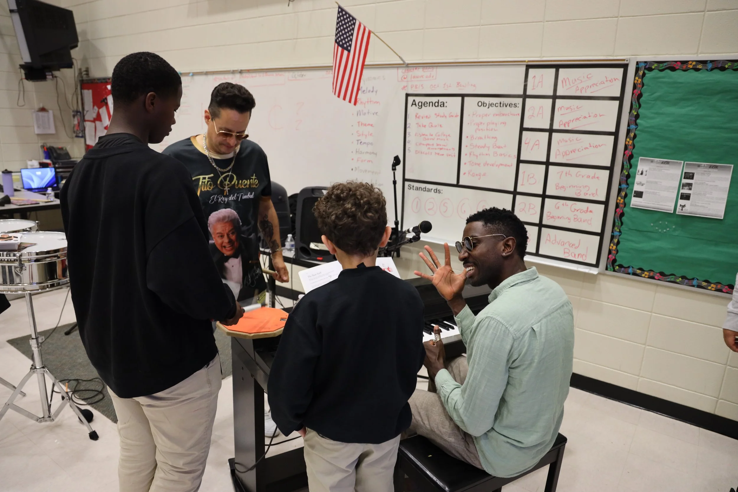 A group of students and a teacher in a music classroom. Four people are visible: a man sitting at a keyboard with sunglasses, a woman standing nearby with a small American flag, a person with curly hair, and another person with short dark hair. The classroom has a whiteboard with an agenda and objectives related to music lesson plans, and musical equipment like drums are visible.