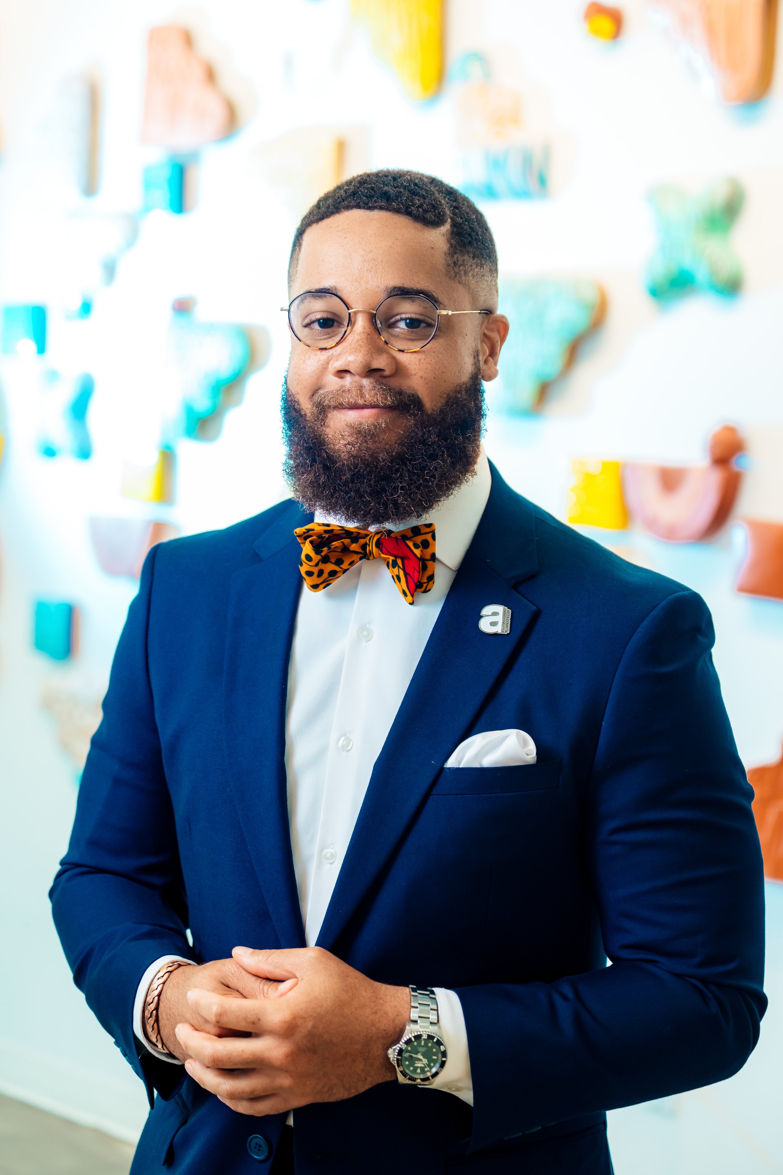 A man in a dark blue suit, white shirt, and colorful bow tie standing in front of an art wall, wearing glasses, a wristwatch, and a lapel pin.