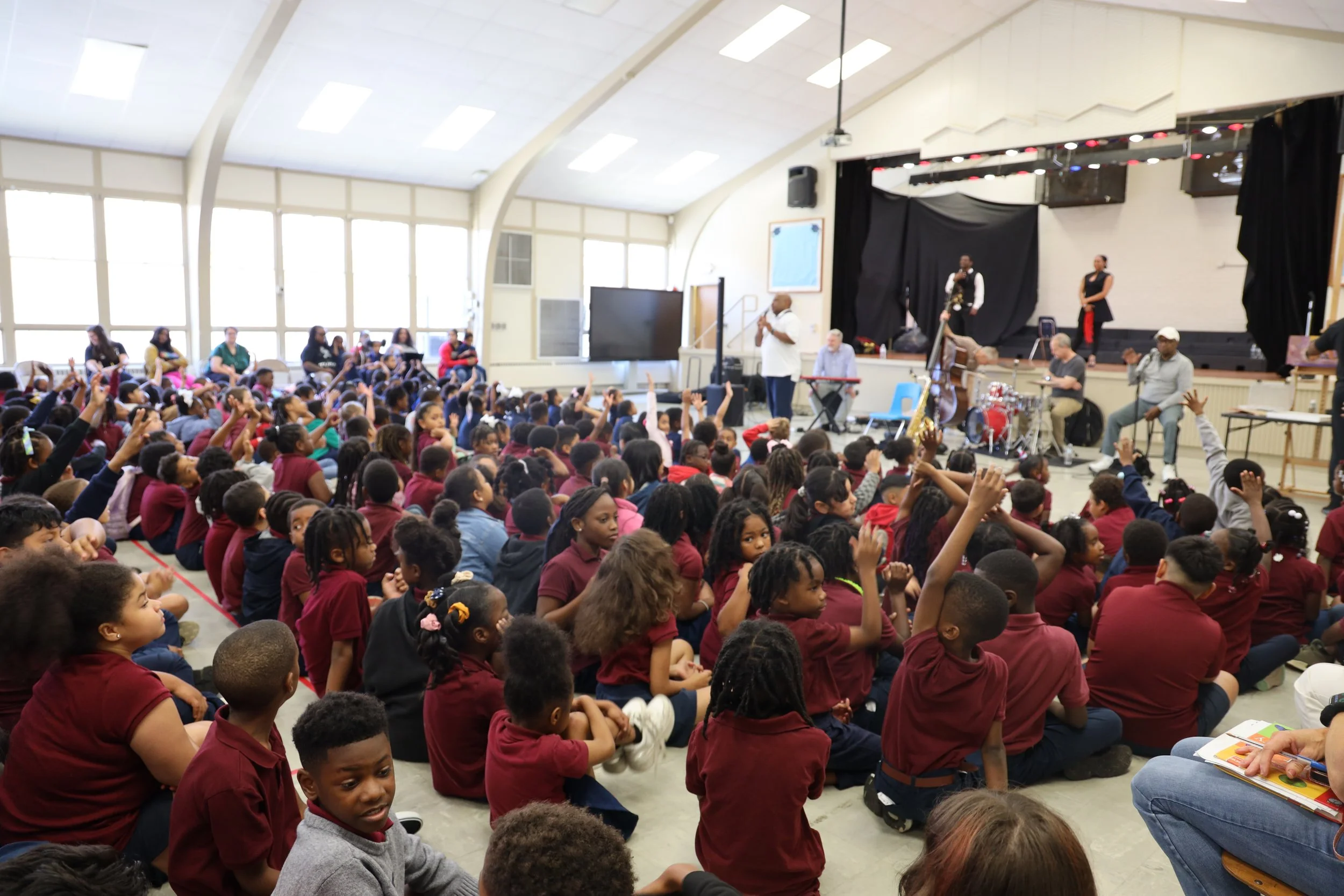 A large group of children seated on the floor of a school gymnasium, attentively listening to a man in a white shirt speaking on stage with musical equipment, while several adults observe from the side.