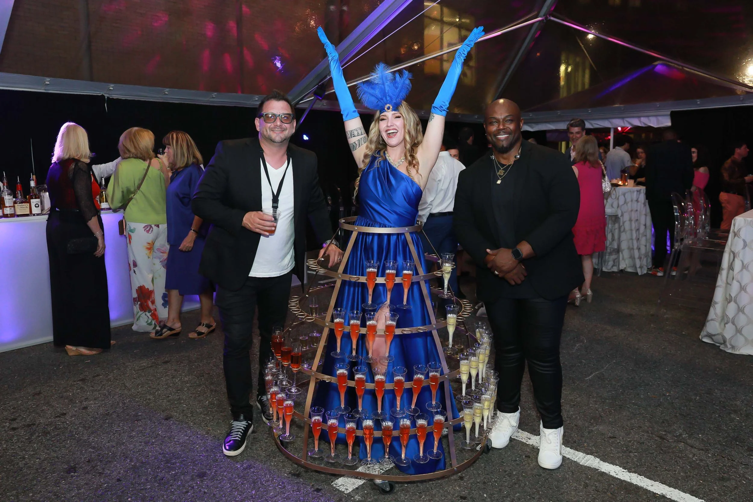 People at a party surrounding a tower with multiple glasses of drinks, with one woman dressed in an elaborate blue costume with gloves and a feathered headpiece in the center, smiling and raising her arms.