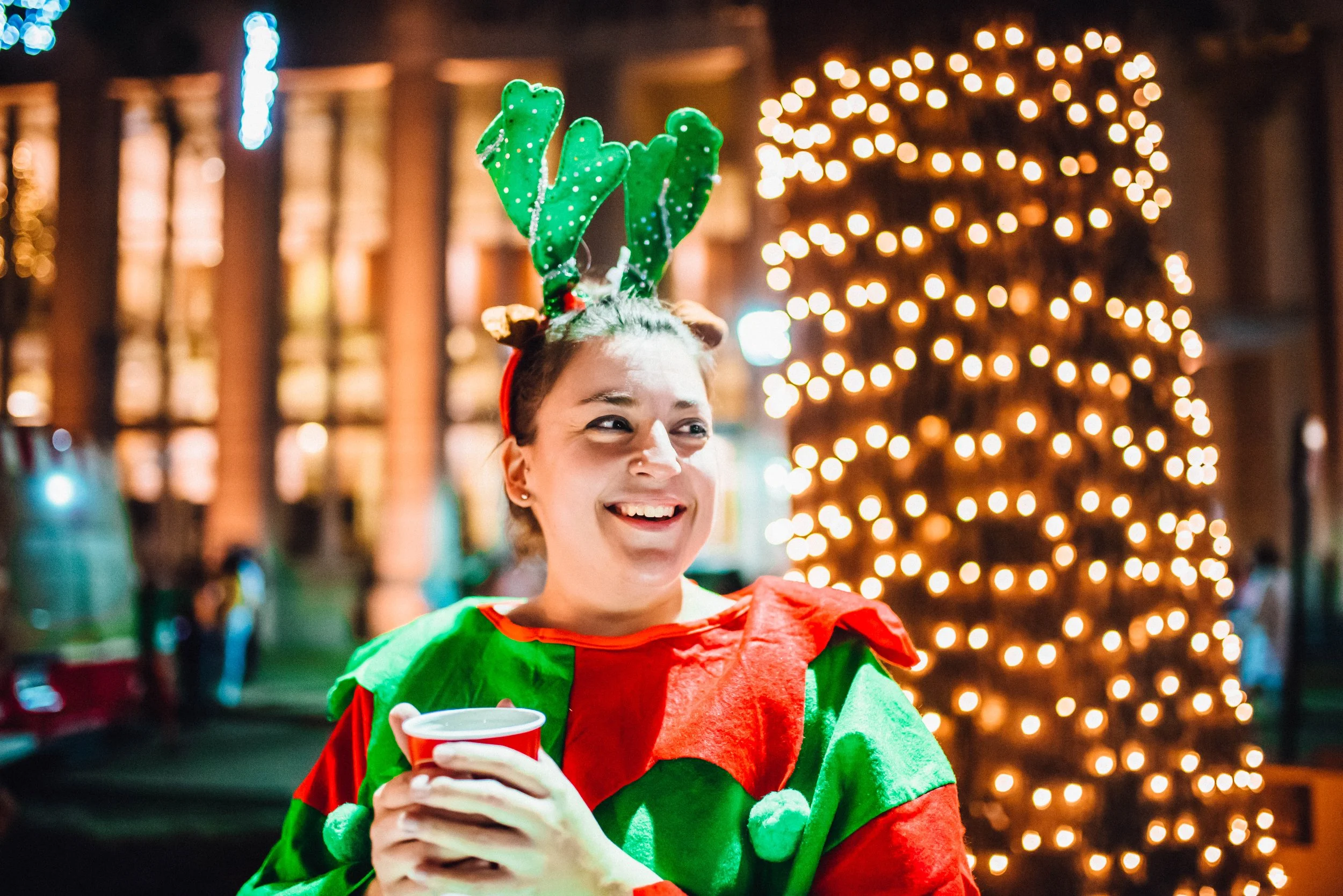 A woman smiling at a Christmas party, wearing a candy cane themed reindeer antlers headband and a red and green festive outfit, holding a red cup, with a background of Christmas lights and decorations.