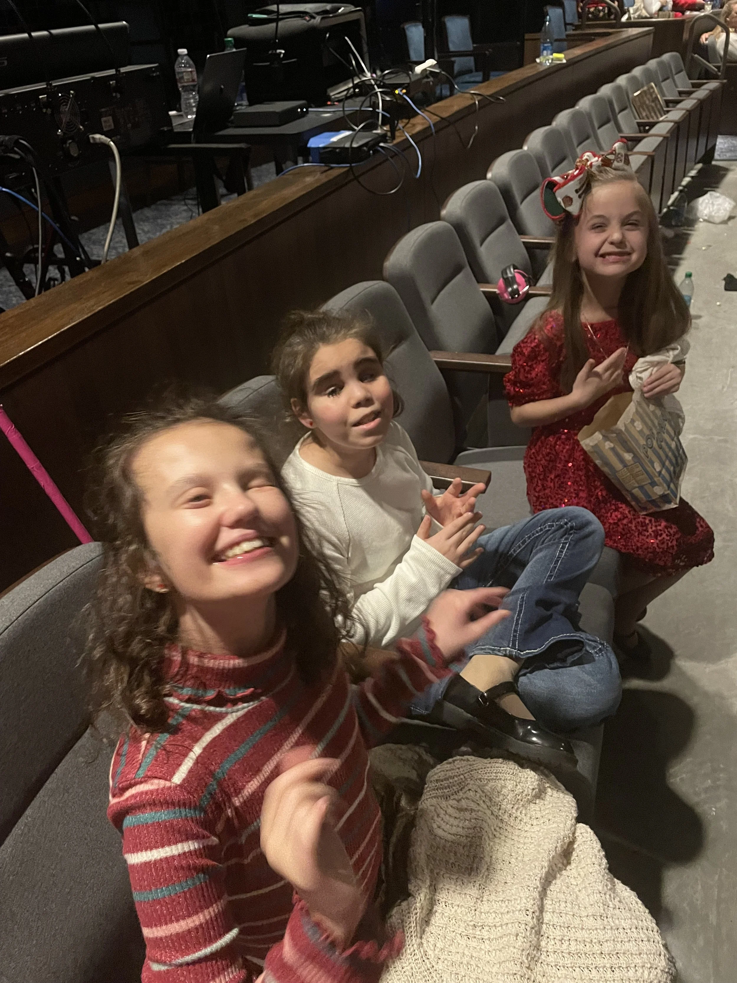 Three young girls sitting in a row at an indoor event, smiling and making facial expressions. The girl on the far right is wearing a red dress with a bow in her hair, holding a striped gift bag. The middle girl is wearing a white sweater, and the girl on the far left, partially visible, has curly hair and is smiling widely.
