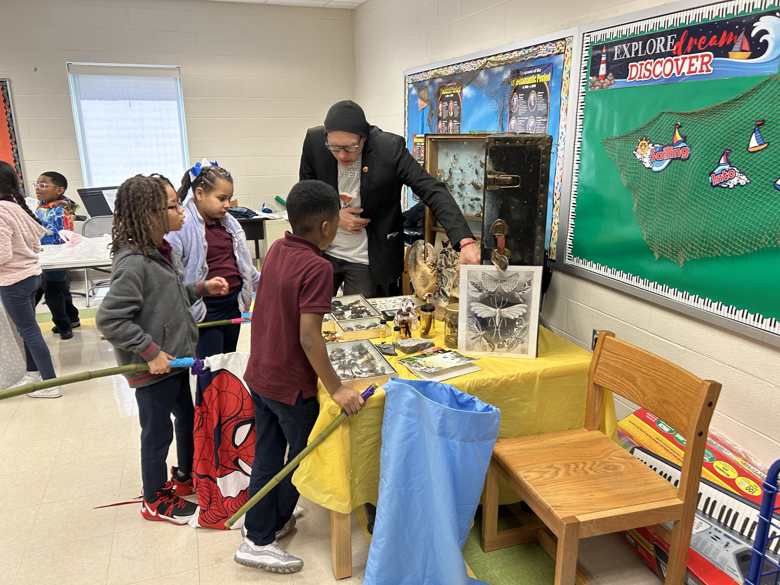 A group of children gather around a table with educational displays and artifacts, in a classroom or library setting, looking at items presented by an adult.