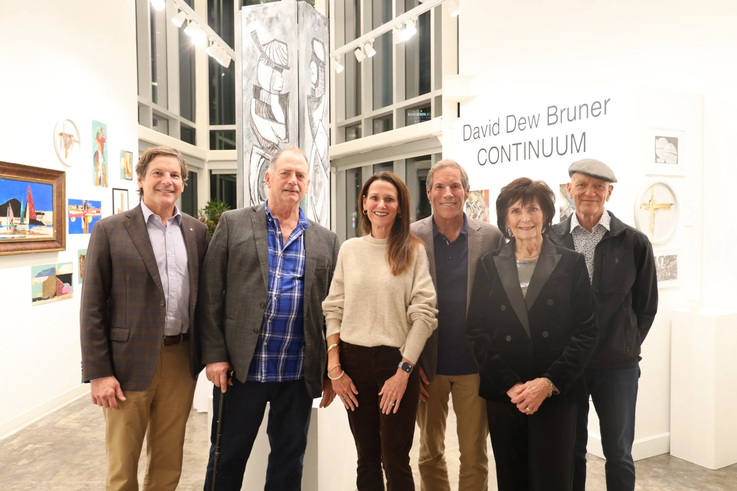 Group of six people standing in an art gallery, smiling at the camera with various artworks displayed on the white walls behind them, and a tall abstract black and white artwork hanging in the center.