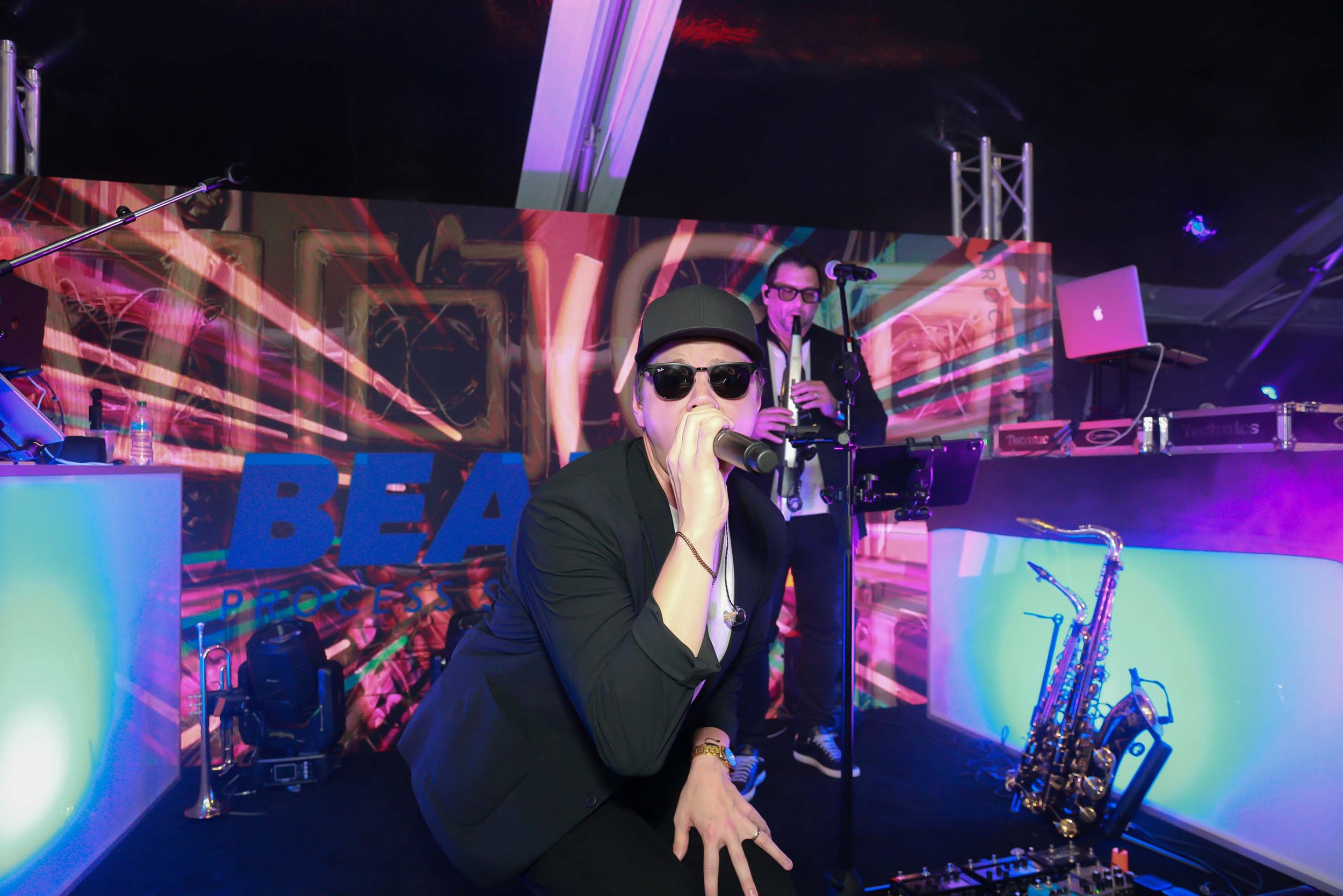 A DJ wearing sunglasses, a cap, and a black jacket, performing at a colorful music event with LED screens and equipment around him.