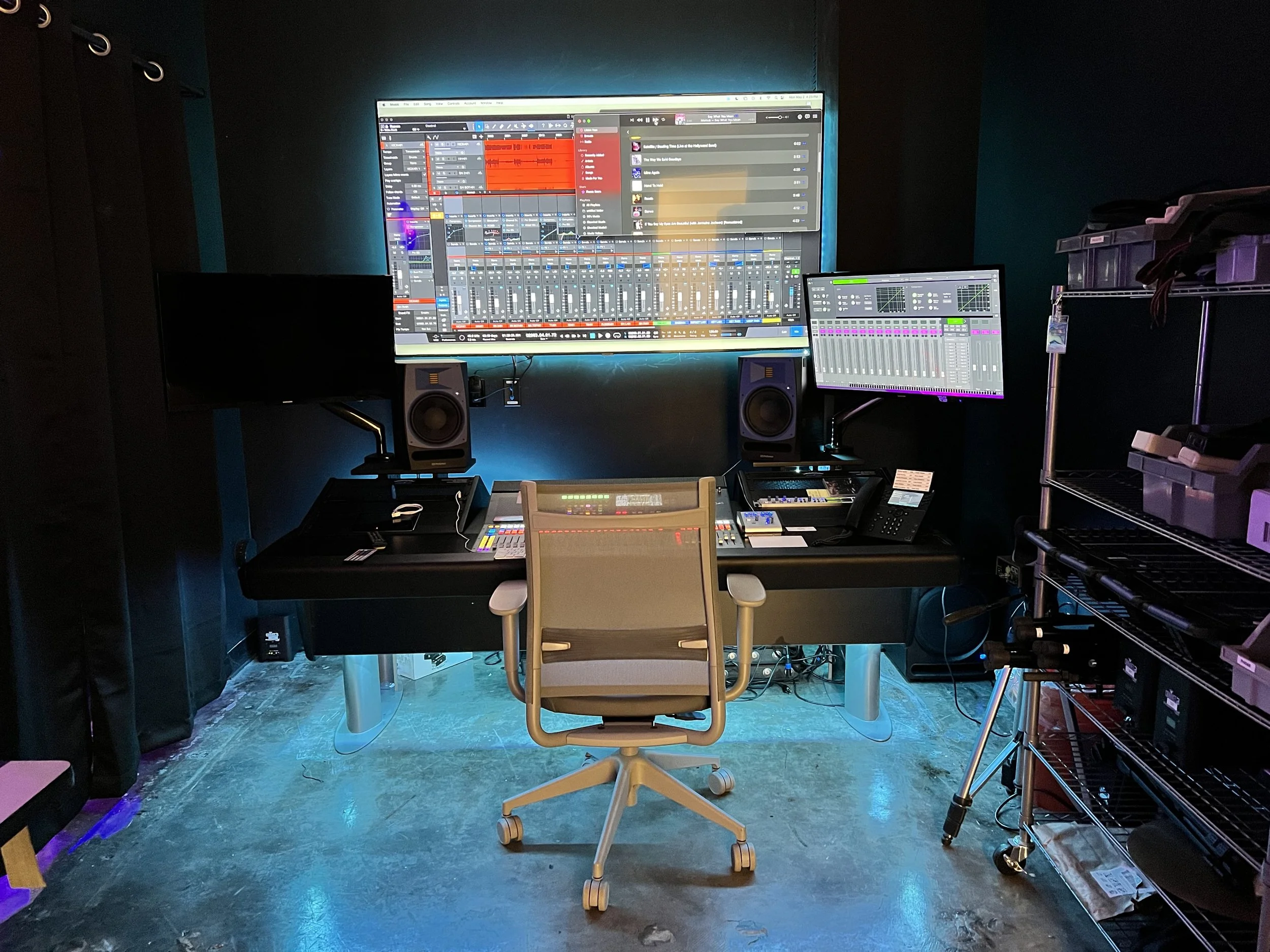 A home music studio with a large wall-mounted monitor displaying digital audio workstation software, smaller monitor, studio speakers, audio equipment on a desk and a chair in front of the setup, lighting with blue tones, and a metal shelf with storage boxes to the right.