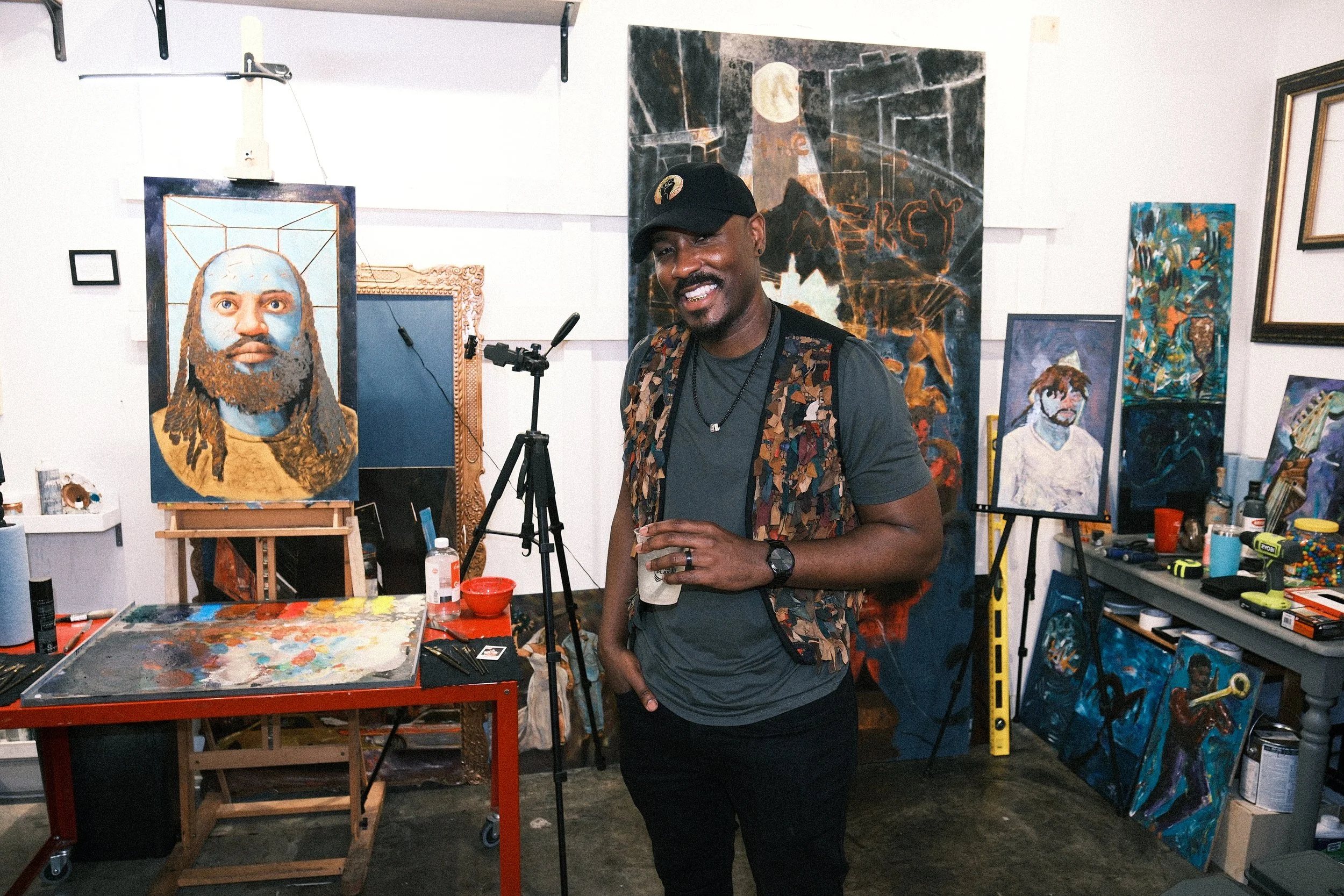 A man smiling inside an art studio, surrounded by various colorful paintings and art supplies. He is wearing a black cap with an emblem, a dark t-shirt, and a vest with a camouflage pattern, holding a drink and standing near a table with paint and art tools.