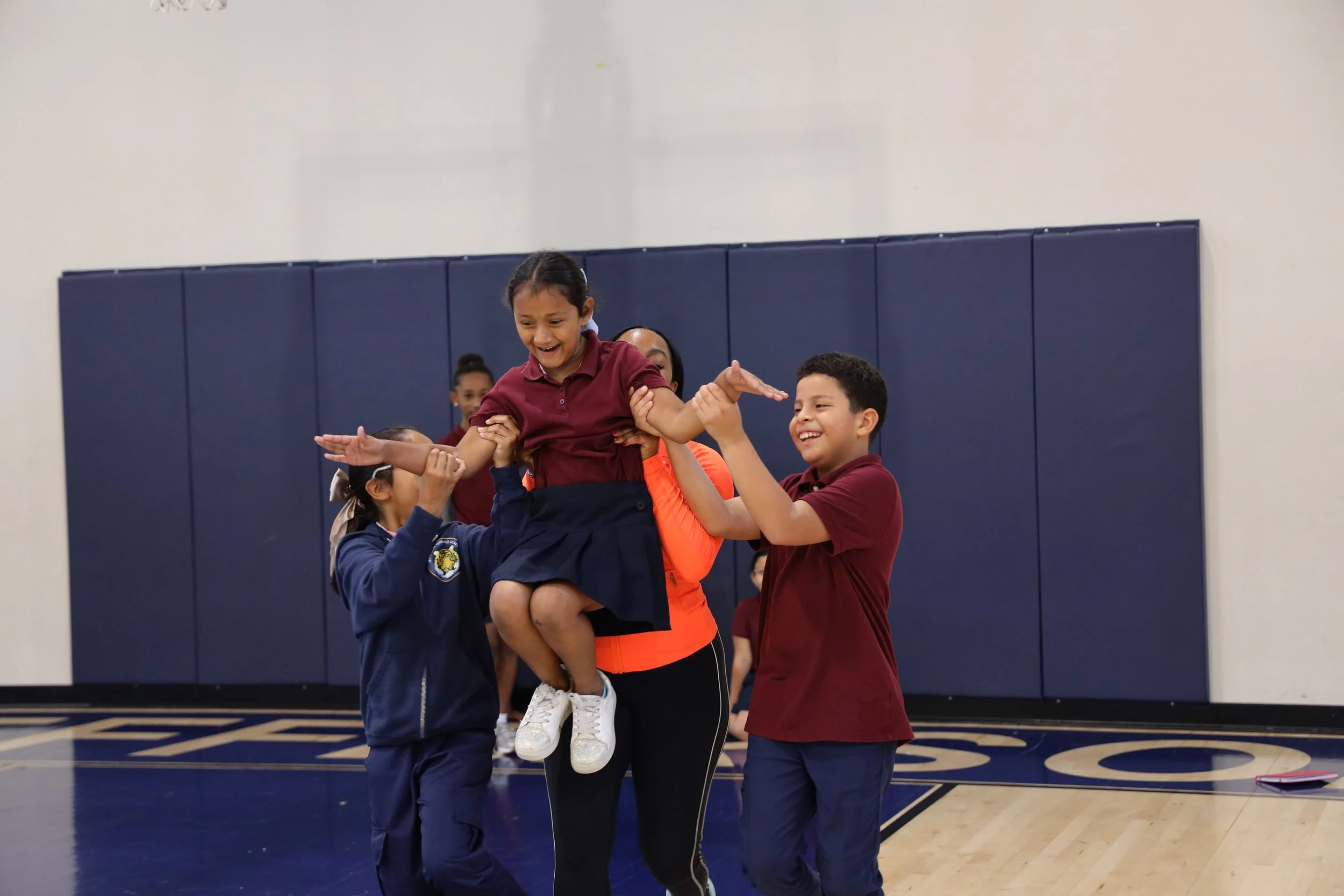 Children and adults in a gymnasium participating in a team-building activity, one girl being lifted by others.