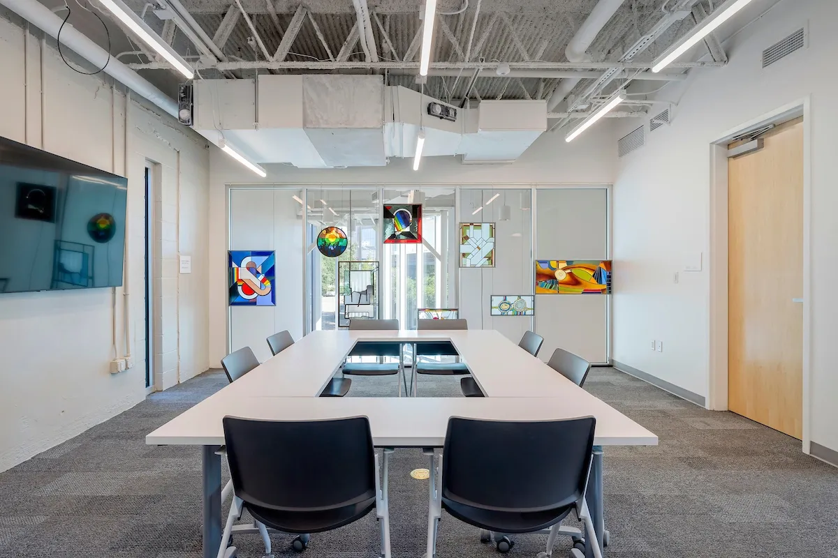 Modern conference room with a rectangular table, black chairs, colorful abstract artwork on walls, large window, and ceiling with exposed ducts and pipes.