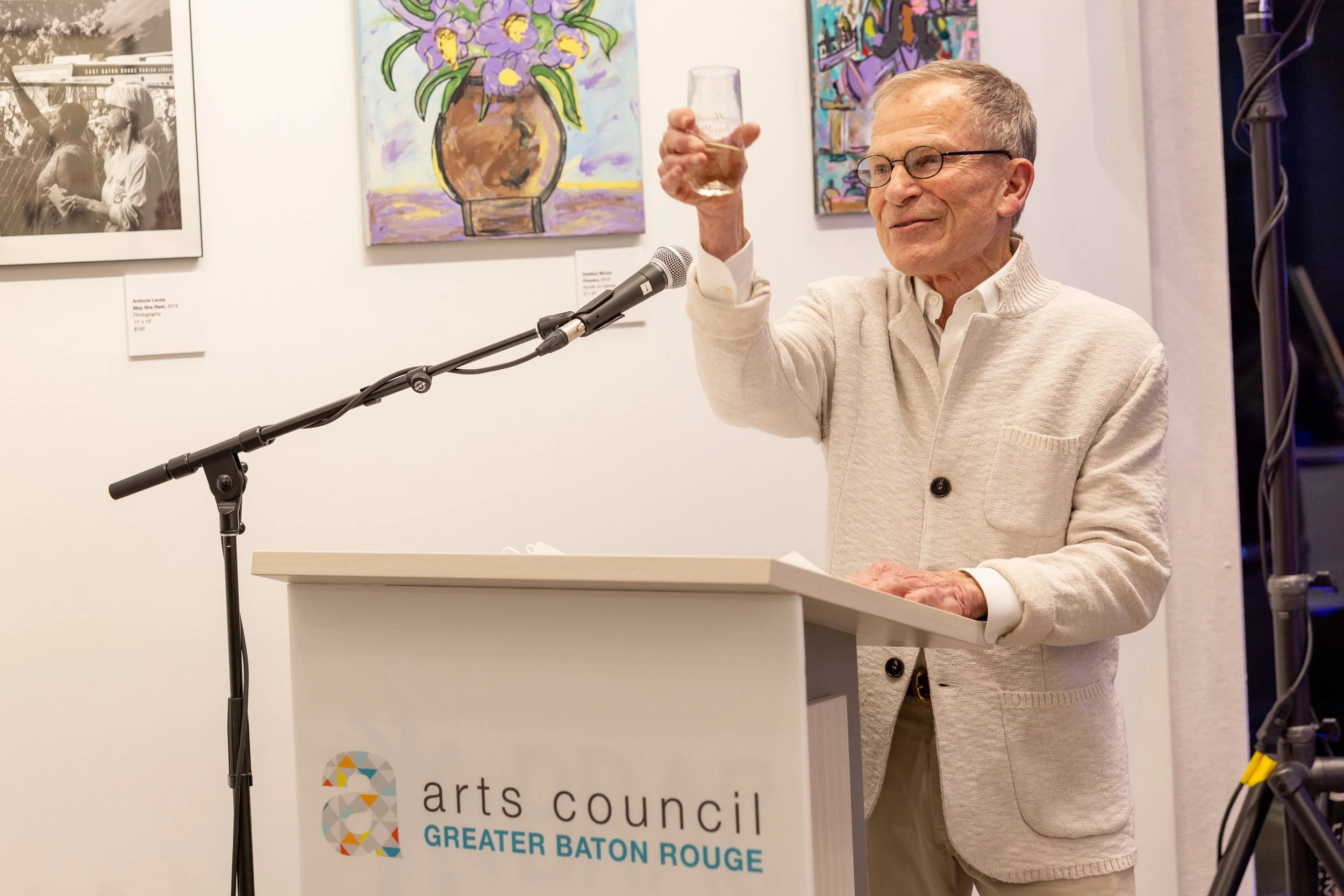 An elderly man standing at a podium with a sign for the arts council, raising a glass in a toast. Behind him are colorful paintings on a wall.